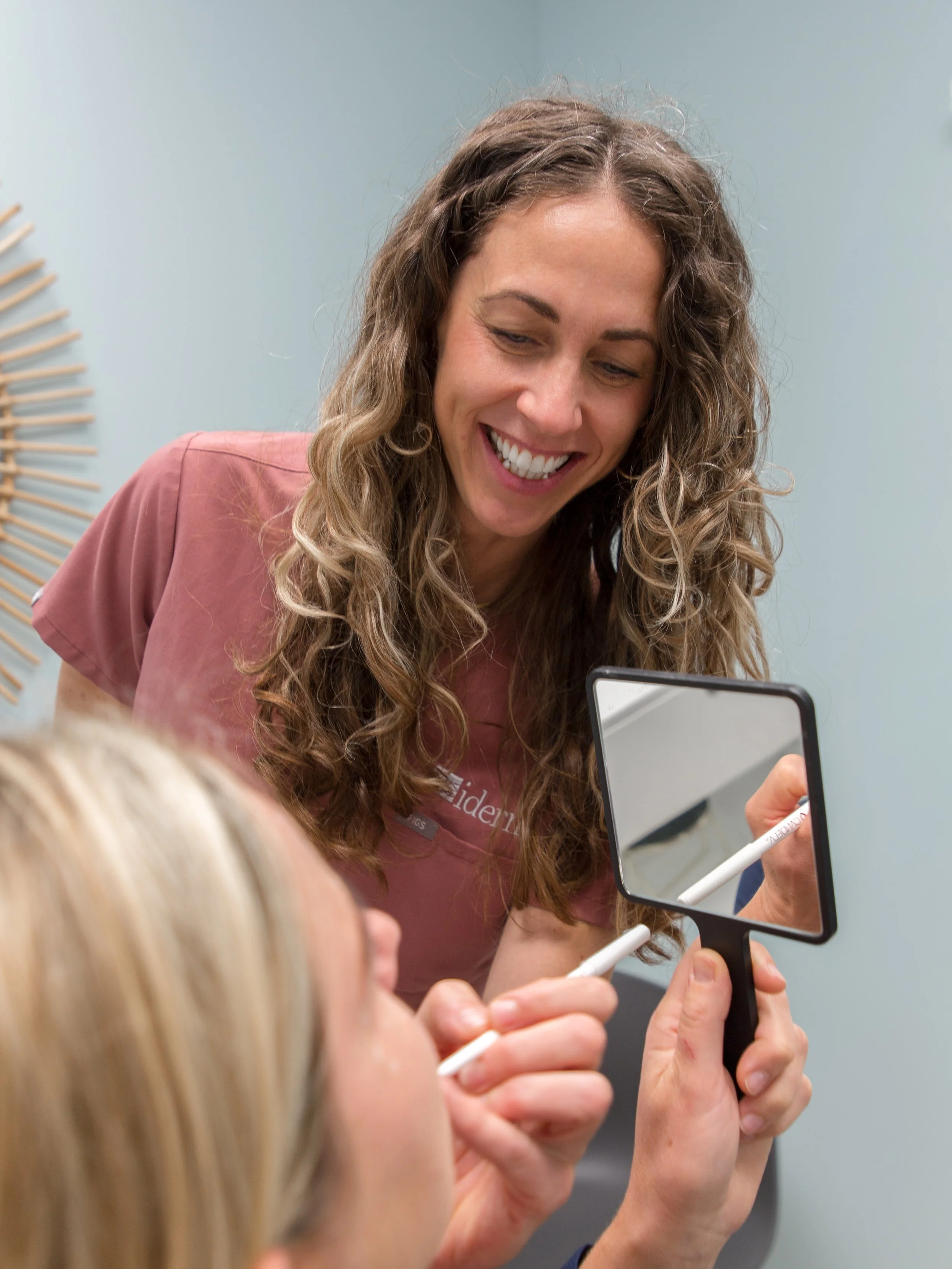 A woman with curly brown hair smiling while holding a mirror and a white marker, engaging with a blonde child, in a light-colored room.