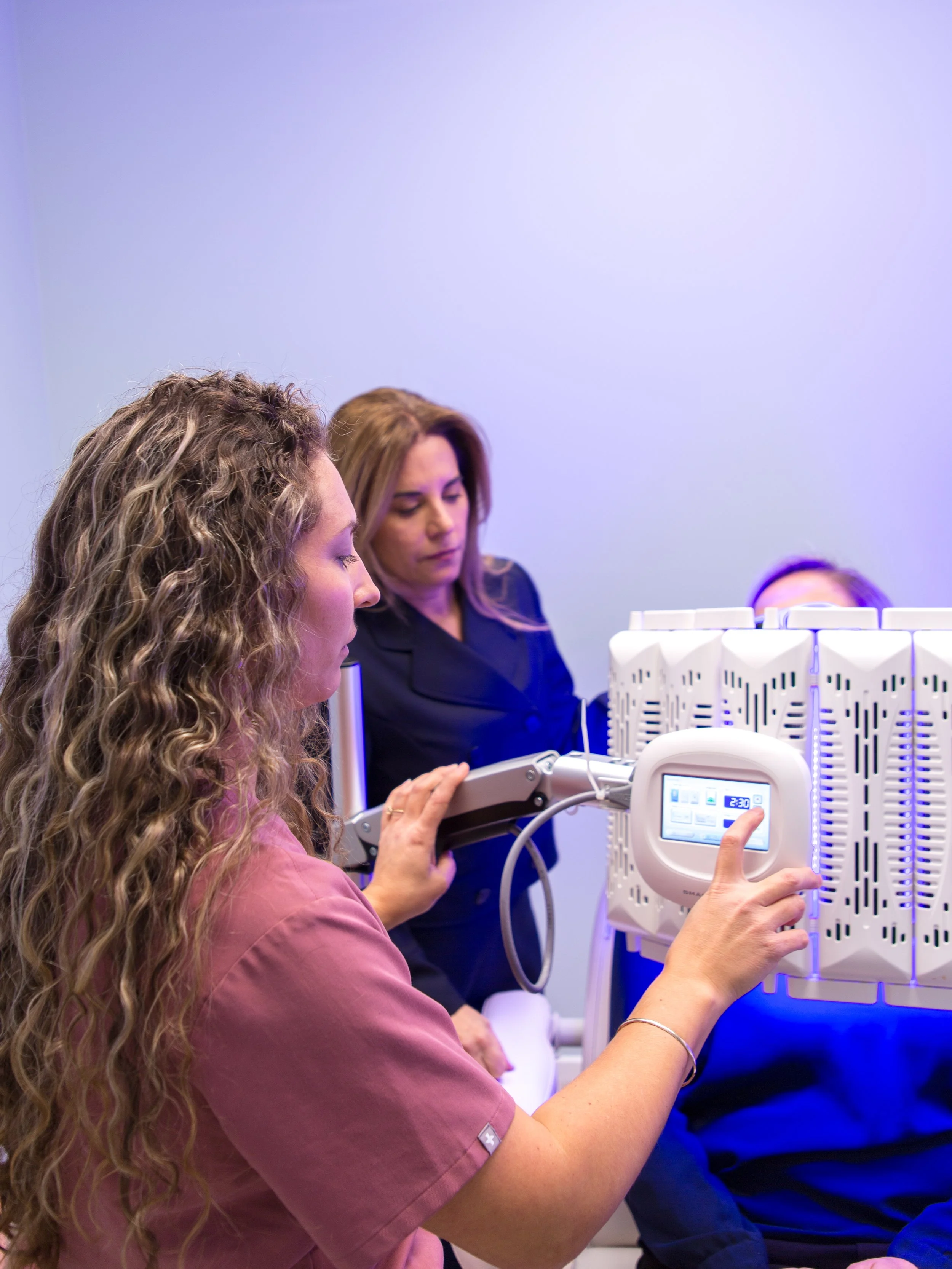 A woman with long, curly hair in a pink uniform adjusts medical equipment while a nurse in blue scrubs observes nearby in a clinical setting.
