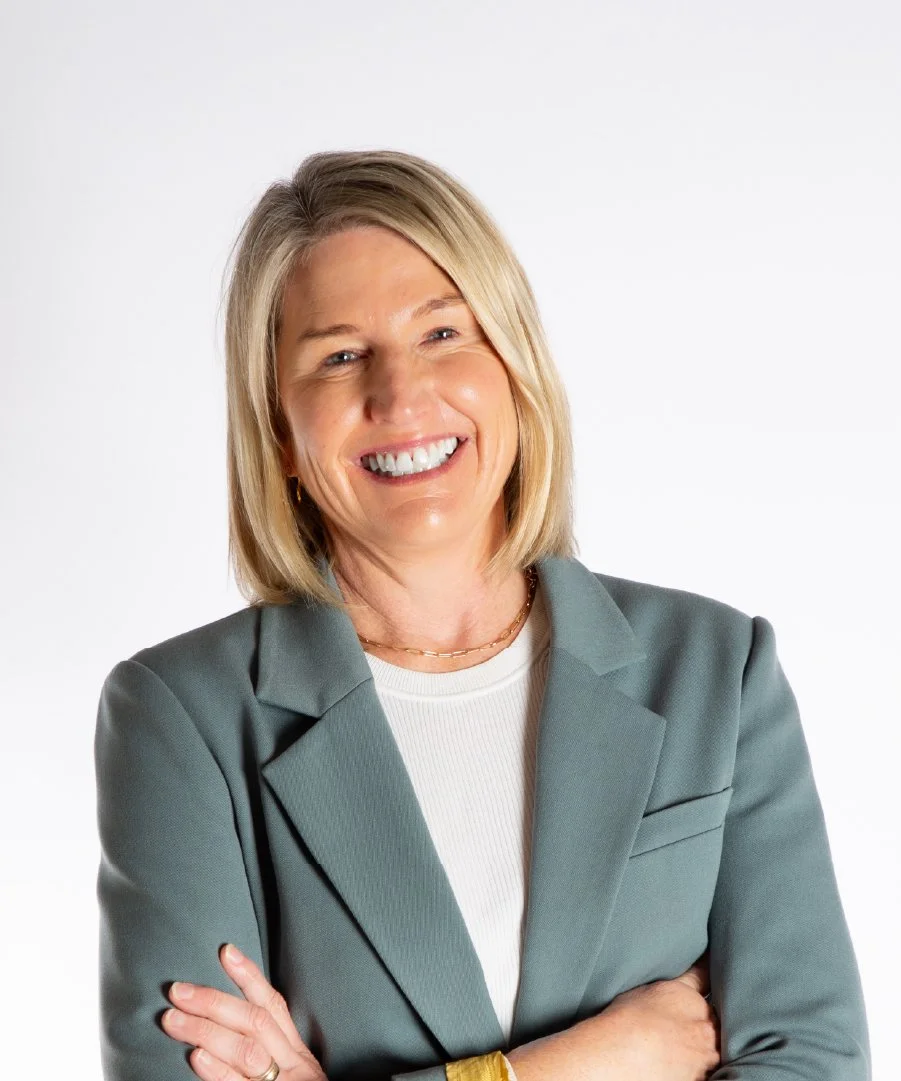 A smiling woman with shoulder-length blonde hair, wearing a light gray blazer and white top, standing with crossed arms against a plain white background.