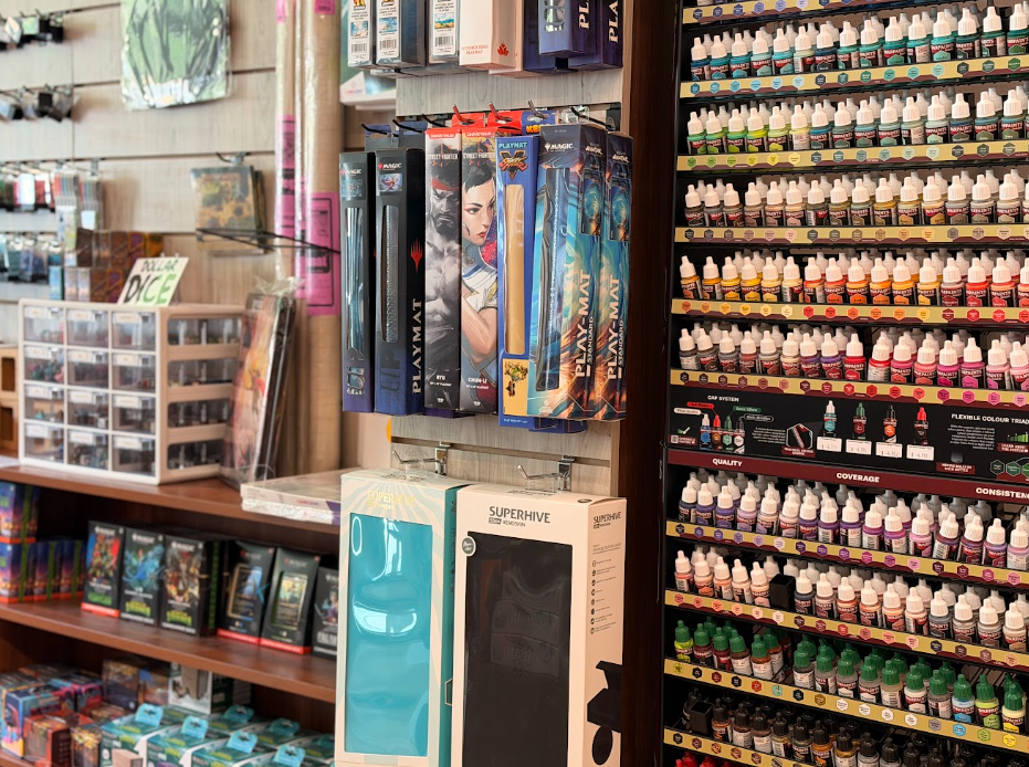 Shelf display of board game boxes, paint bottles, and gaming accessories in a retail store.