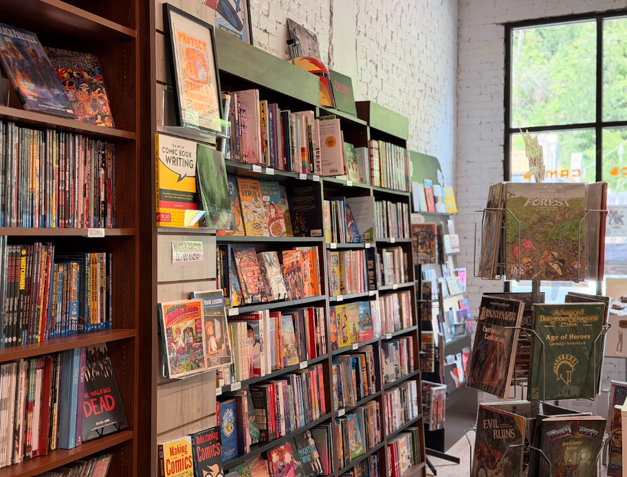 Bookshelves filled with comic books and graphic novels inside a bookstore. A large window letting in natural light is visible in the background.