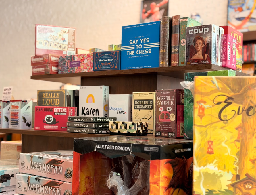 Books on a wooden shelf with titles like "Say Yes to the Chess," "Horrible Couple," "Really Loud," and a game titled "Adult Red Dragon." Some boxed items, including a box with a dragon illustration labeled "Adult Red Dragon," and a box of "Exploding Kittens."