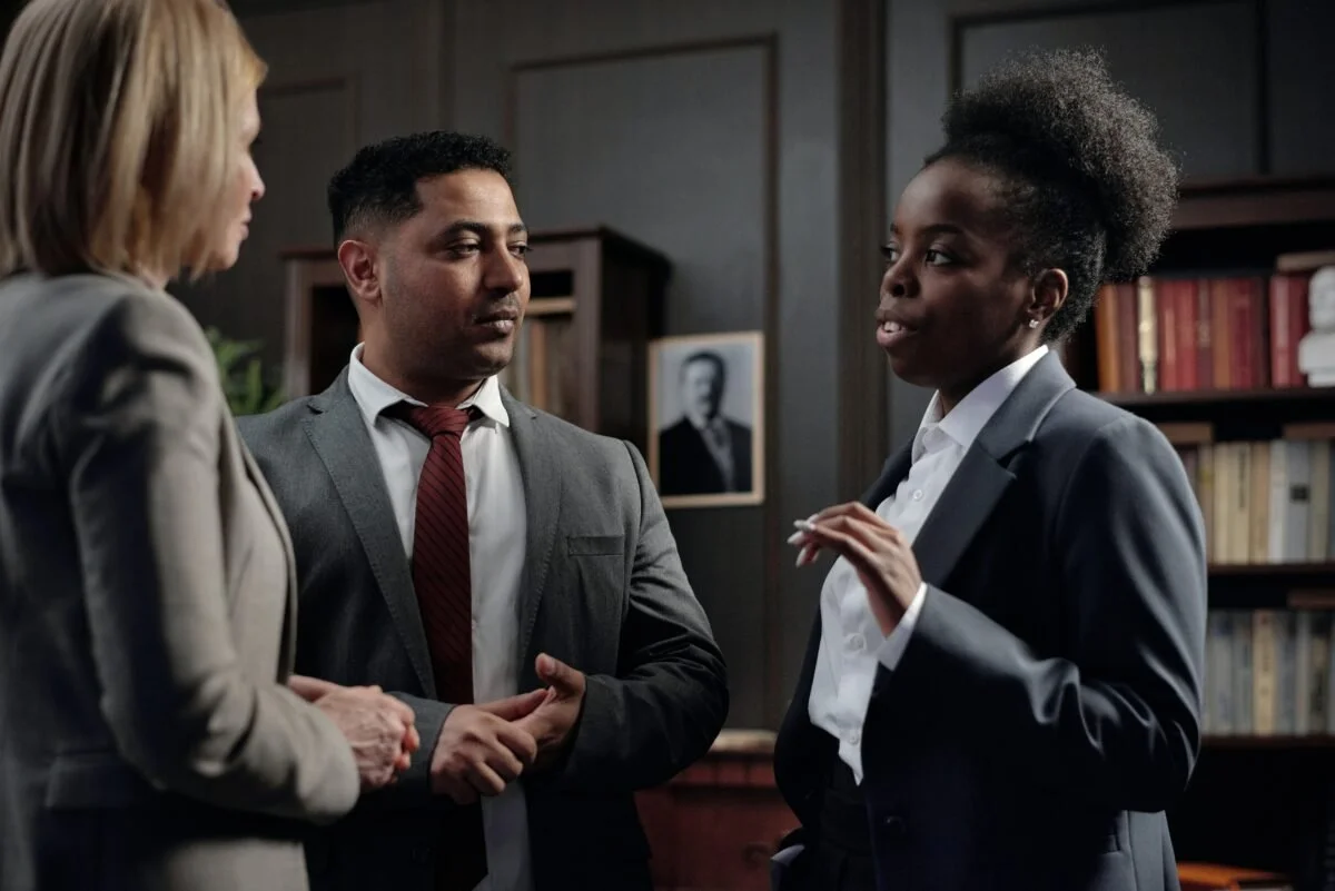 Three professionals have a serious conversation in an office or library setting with bookshelves and a framed photo in the background.
