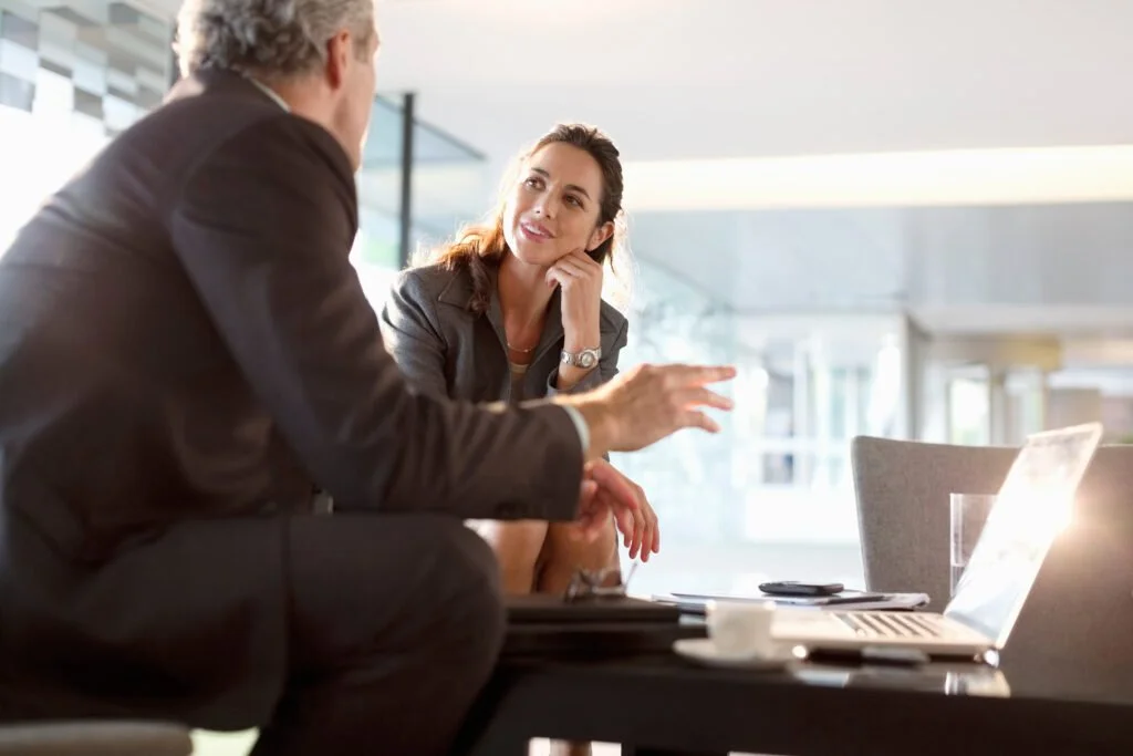 A woman and a man having a conversation in a modern office setting with large windows. The woman is listening attentively while the man is gesturing with his hand.