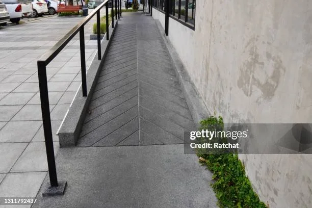 Sidewalk with a ramp and black handrails along a building wall, with parked cars visible in the background.