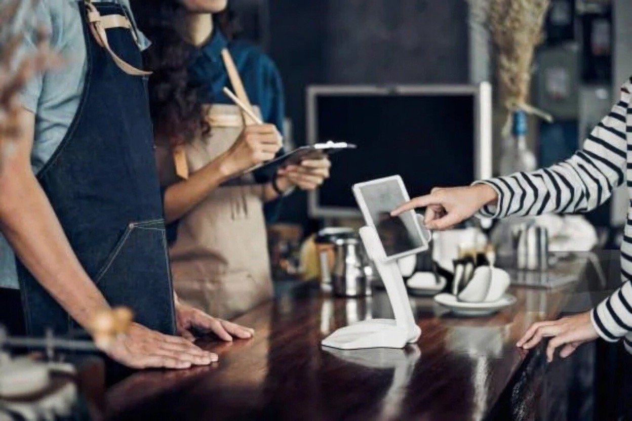 Customer paying at café counter with a tablet POS system, barista taking order in the background.