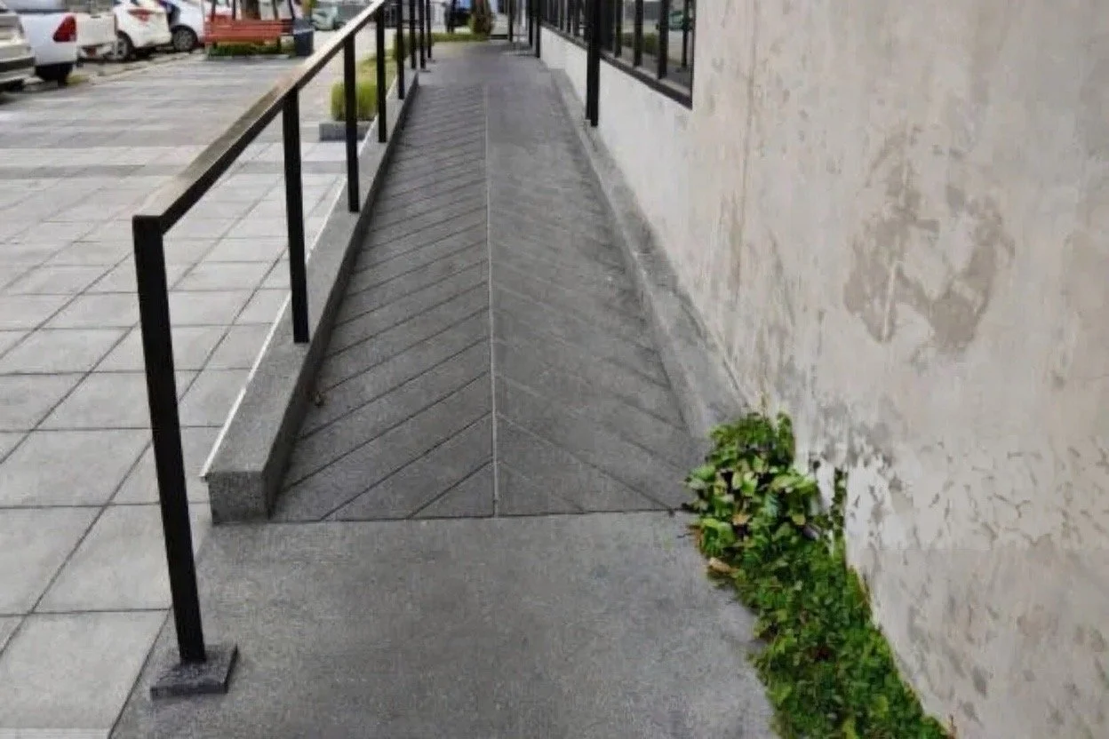 Sidewalk with a ramp and black handrails along a building wall, with parked cars visible in the background.