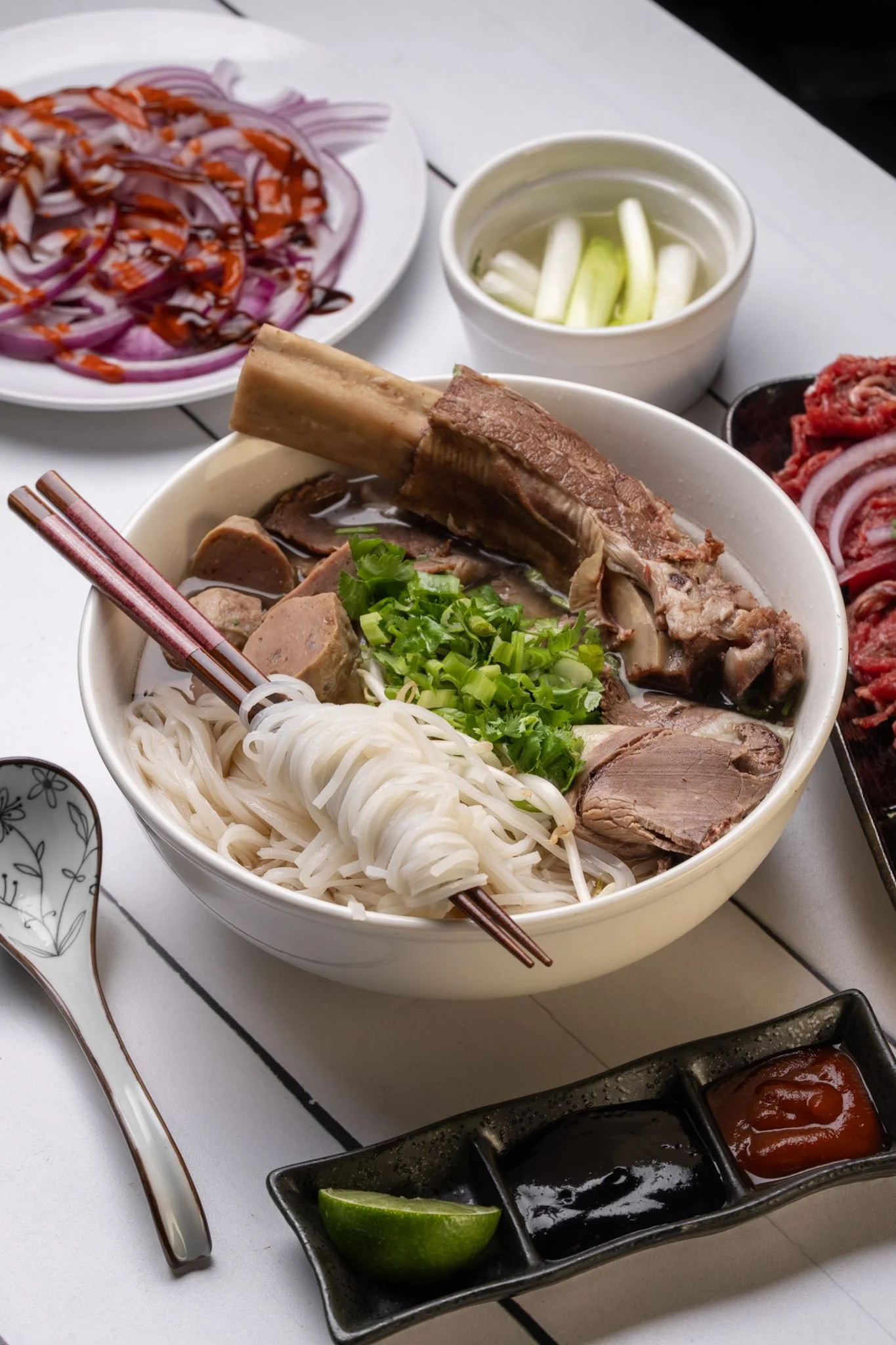 A bowl of Vietnamese pho with beef, noodles, and green onions, accompanied by lime and various condiments, on a white table.