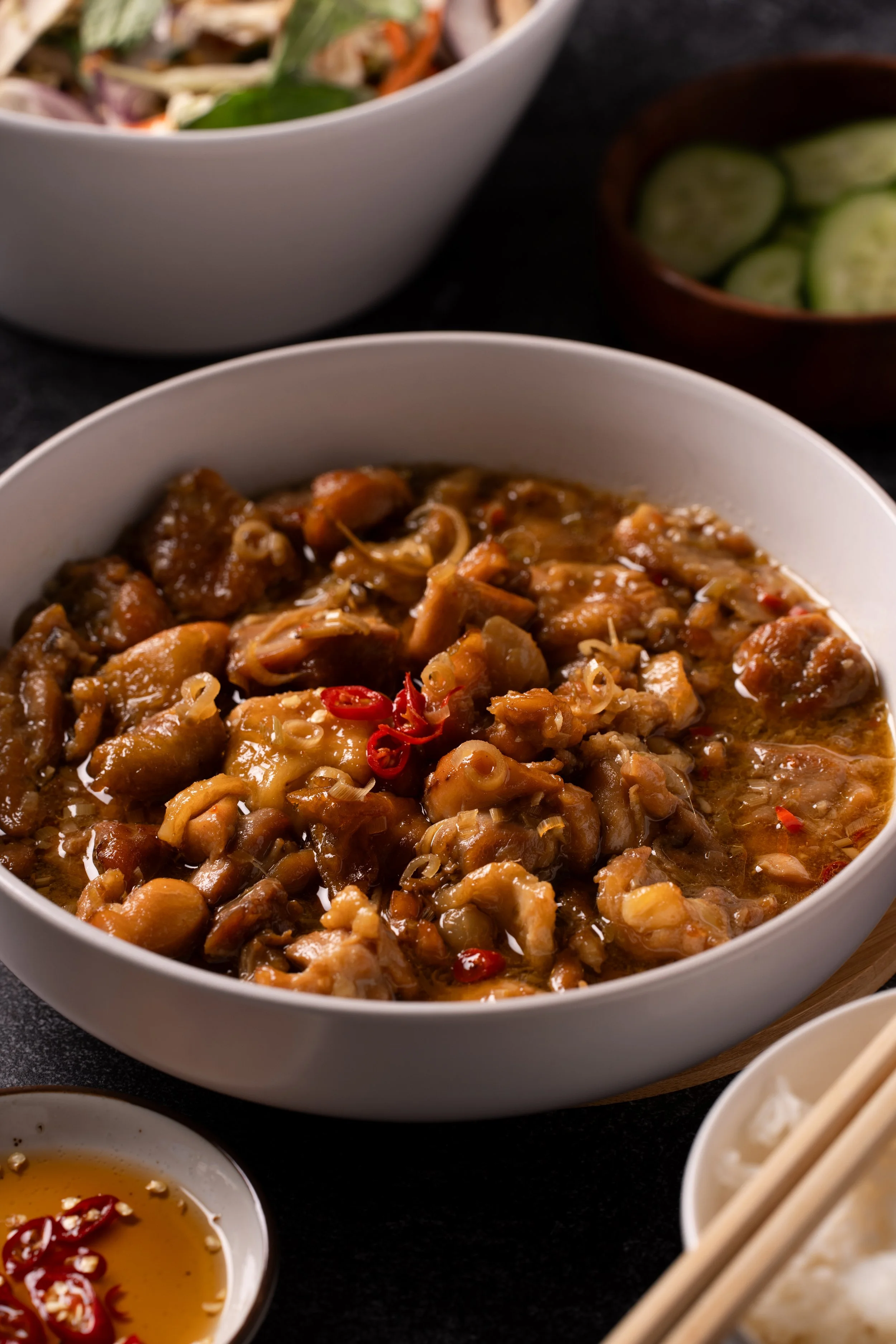A bowl of savory chicken and vegetable stew garnished with sliced red chili peppers, with side dishes of sliced cucumbers in a wooden bowl and a small bowl of dipping sauce with sliced chili peppers, rice with chopsticks, and a salad in the backgroun