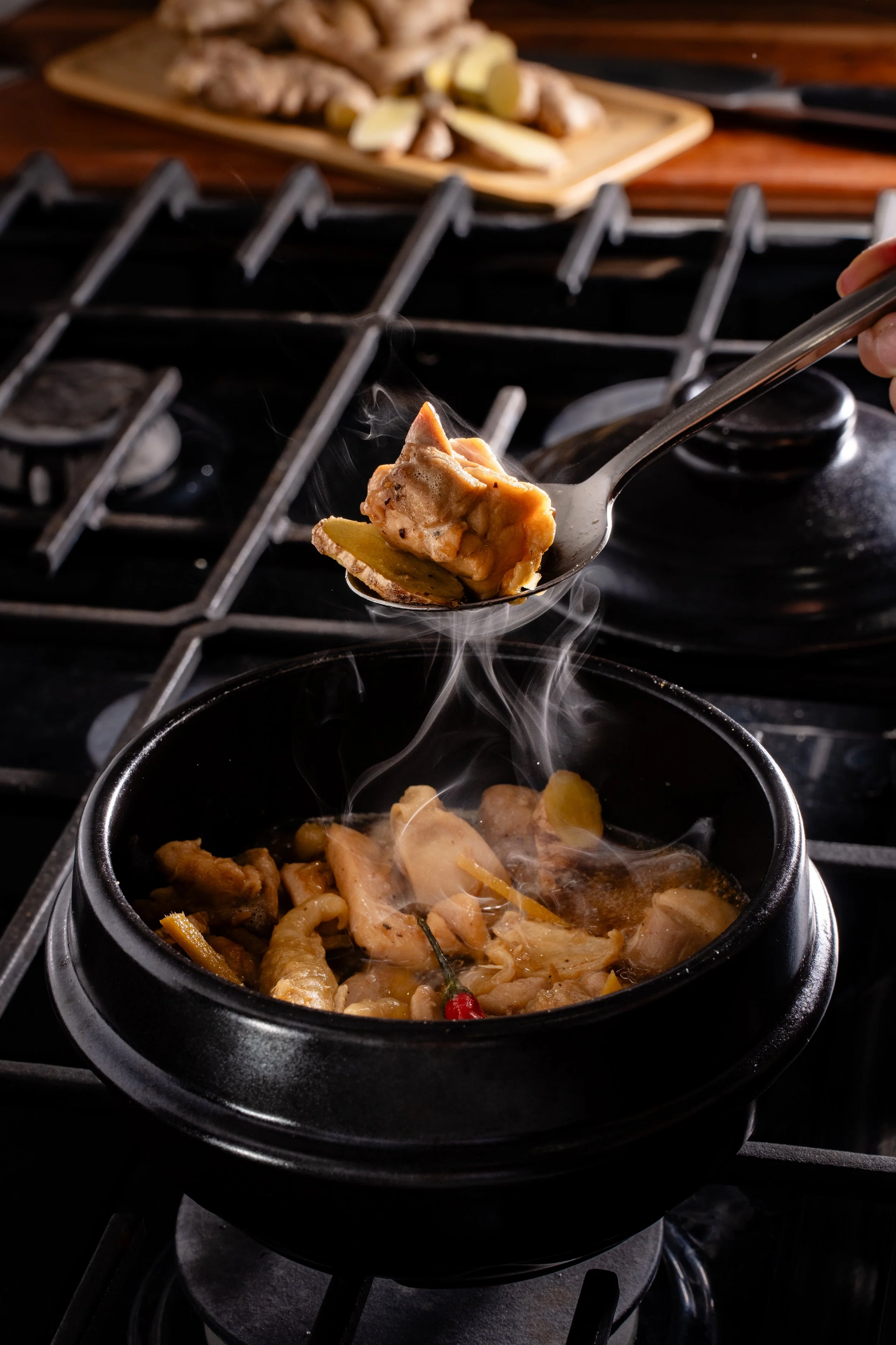 A spoon is scooping ginger braised chicken from a black hot stone bowl on a stove, with a wooden cutting board with ginger in the background.