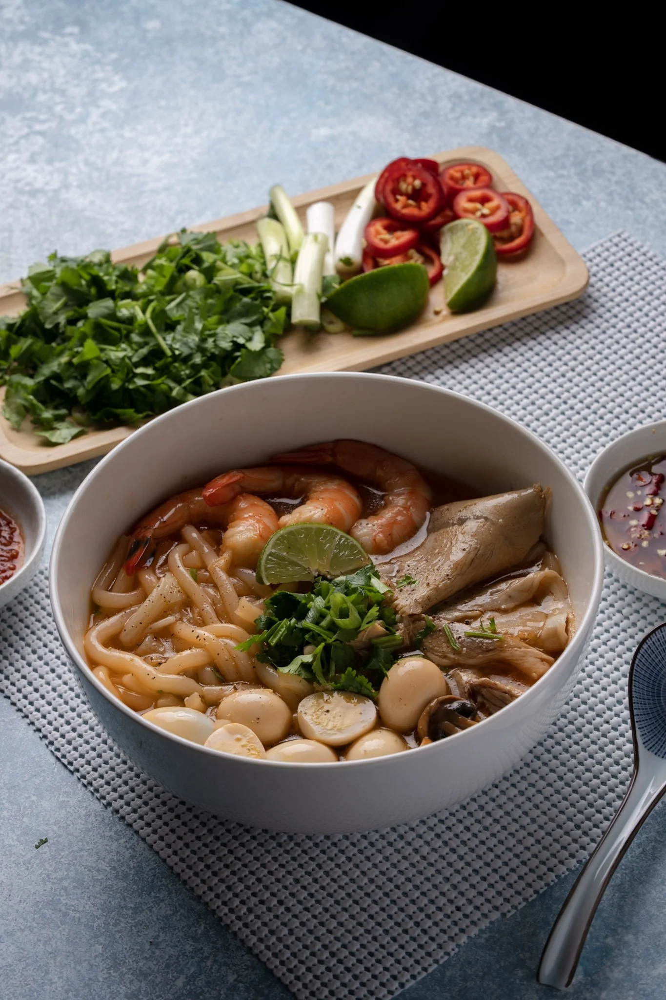 A bowl of Banh Canh with shrimp, quail eggs, and chopped green onions, garnished with lime and cilantro, on a textured tablecloth, with a side of fresh vegetables and dipping sauces.