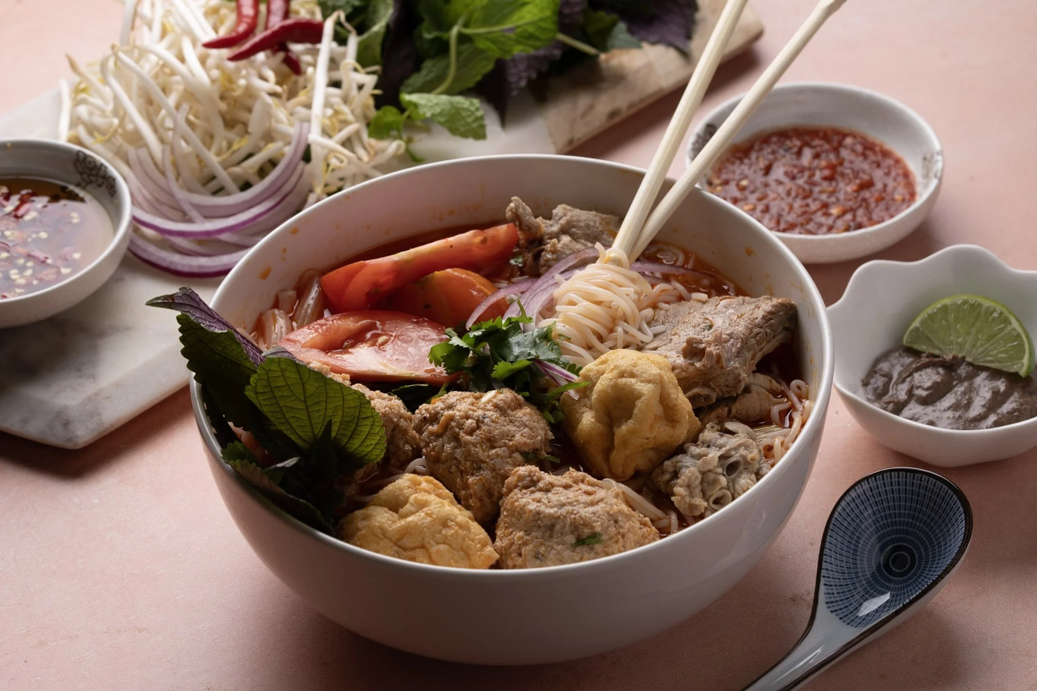 A bowl of Bun Rieu, fresh herbs, and vegetables, accompanied by small dishes of sauces, lime, and bean sprouts on a pink table.