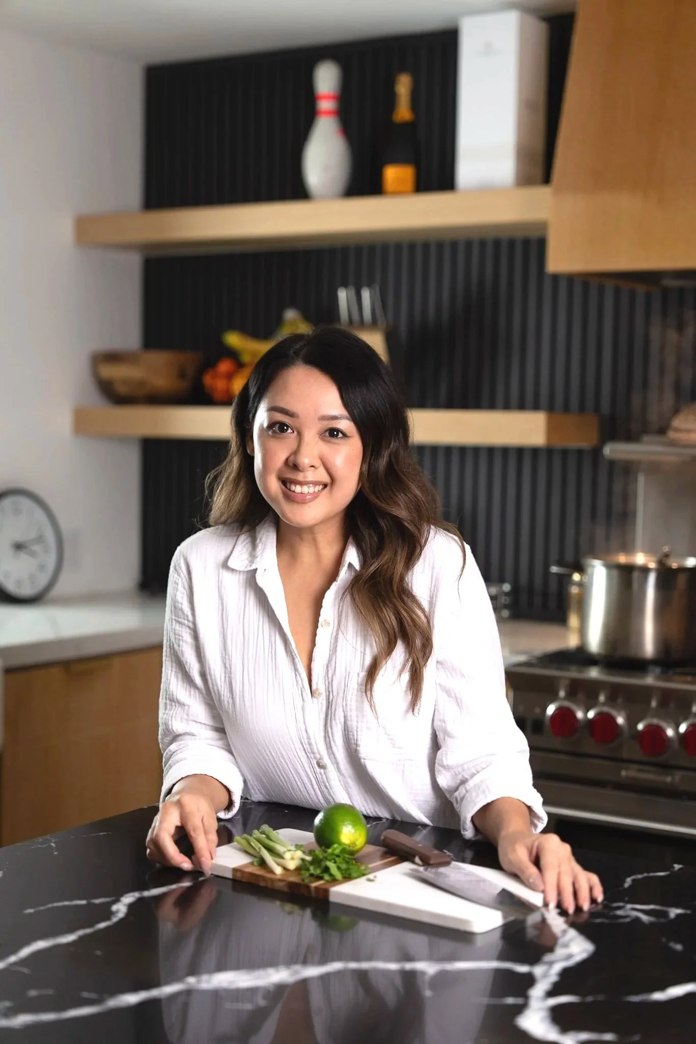 Woman in white shirt smiling at camera in kitchen with a cutting board, lime, green onion, and herbs on the counter.