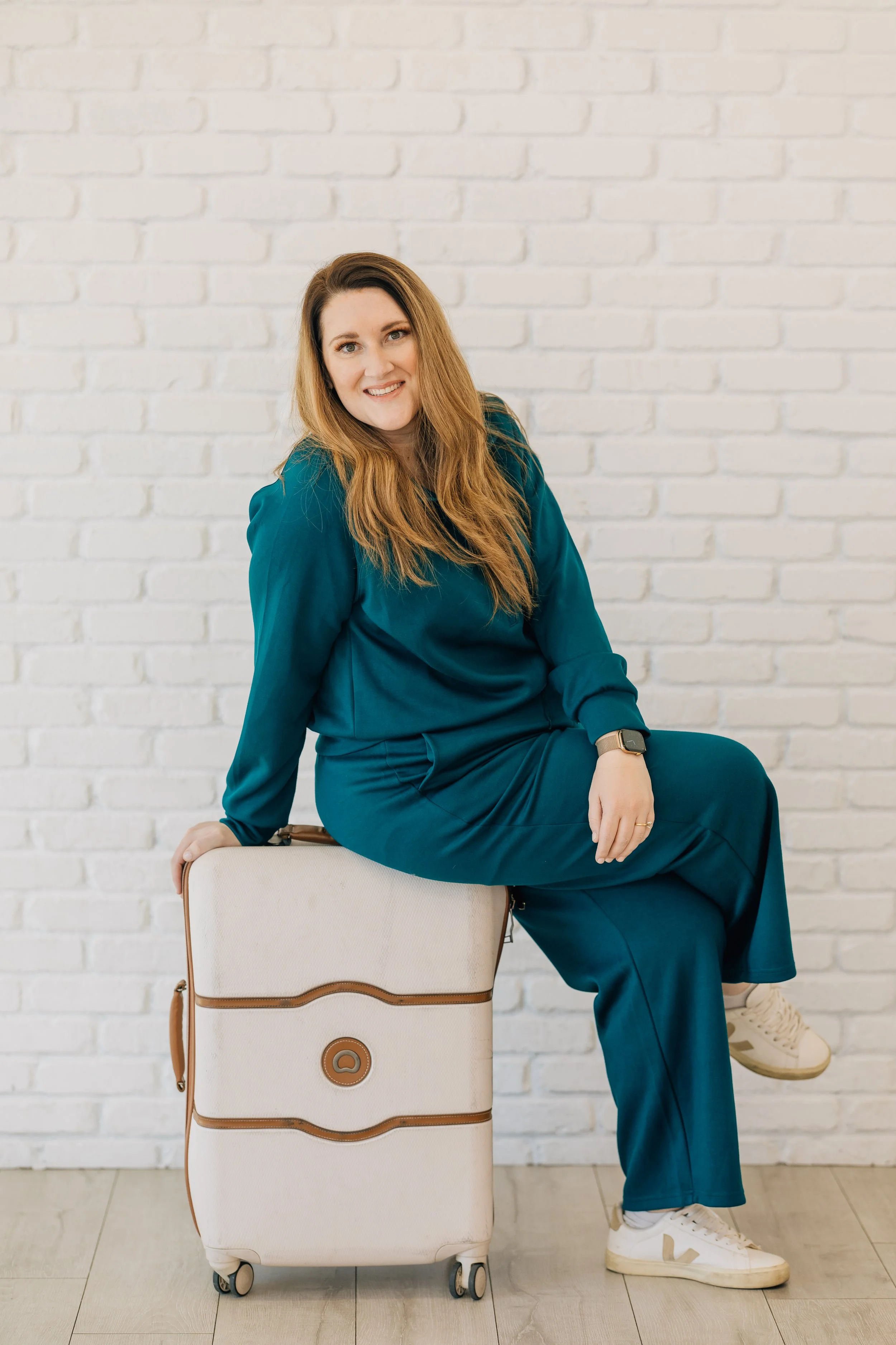 A woman in a teal matching set sits on the edge of a white hardshell suitcase with tan accents.
