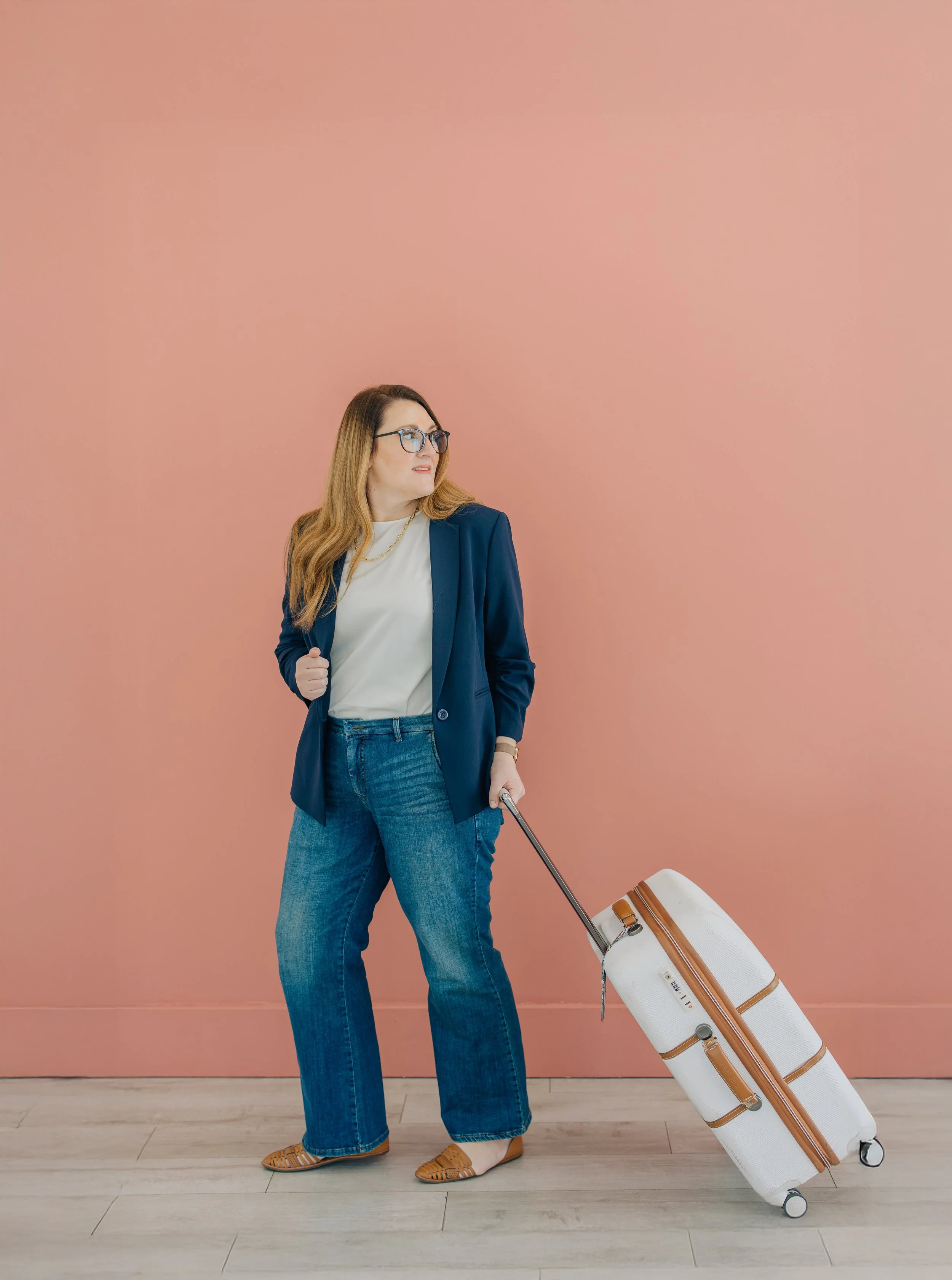 Woman with glasses and blonde hair standing against a pink wall, holding a rolling suitcase, dressed in a blue blazer, white shirt, blue jeans, and tan shoes.
