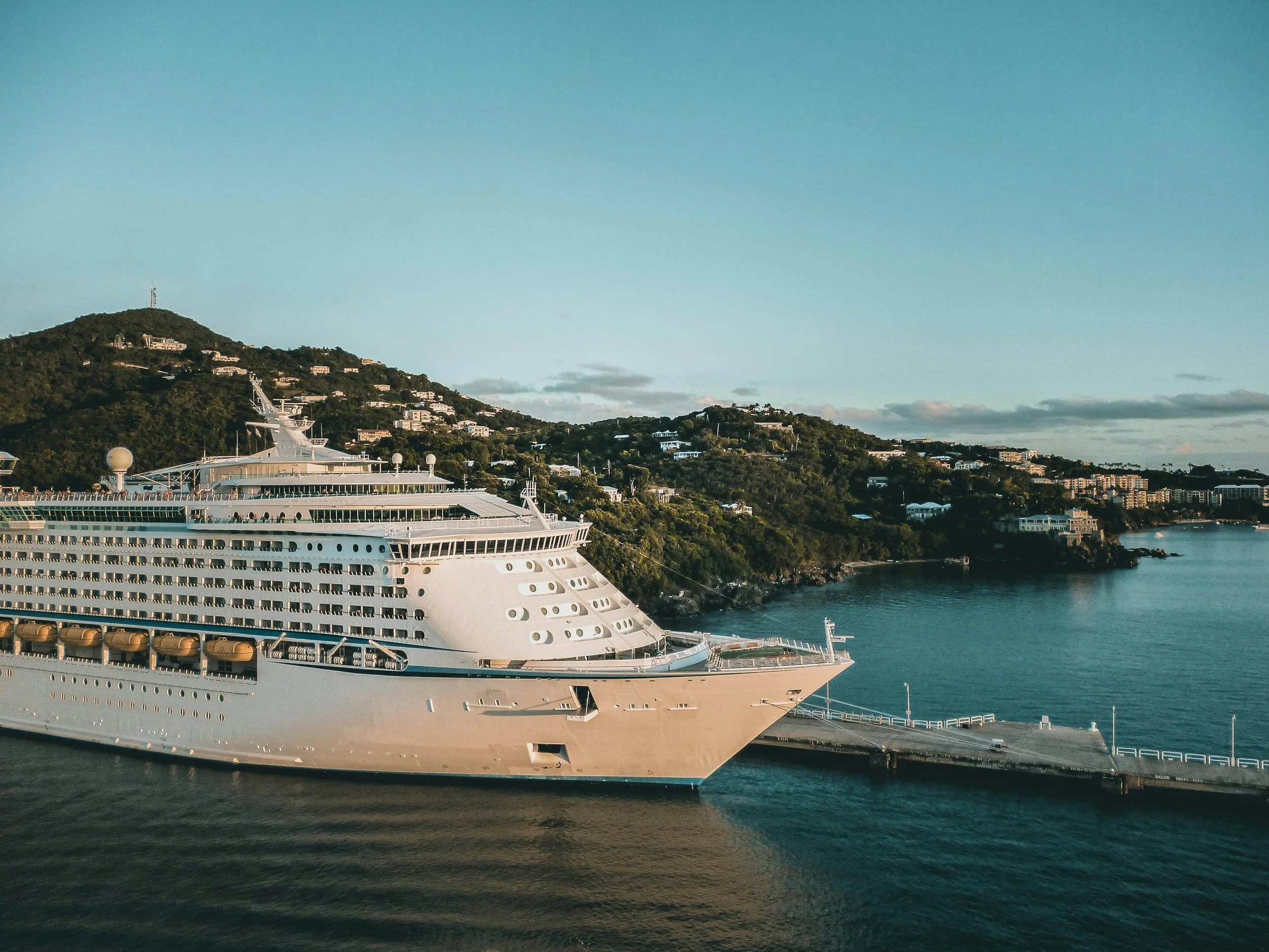 Large white cruise ship docked at a pier with lush green hills and residential buildings in the background under a clear blue sky.