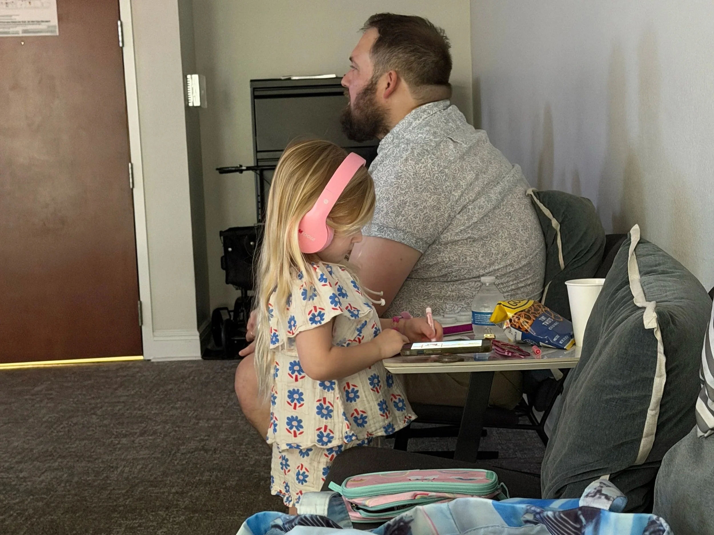 A young girl is watching a show on a phone and coloring while her dad listens to a timeshare presentation.