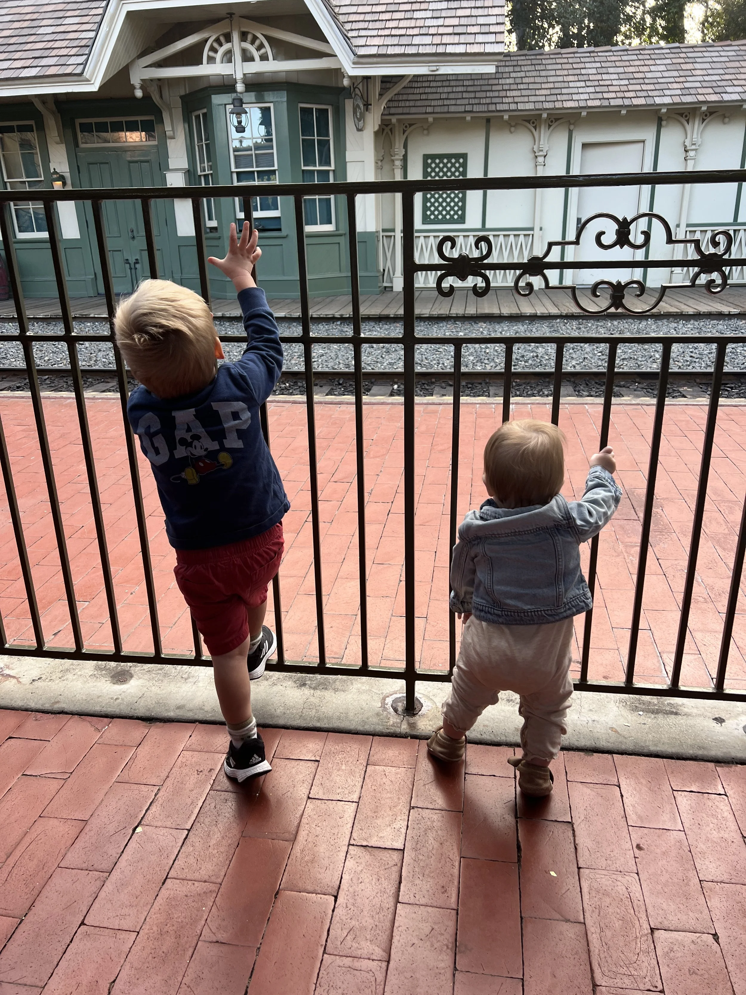 Two young children standing on a brick sidewalk behind a black metal fence, looking and reaching through the bars at a building and a train tracks in the background.