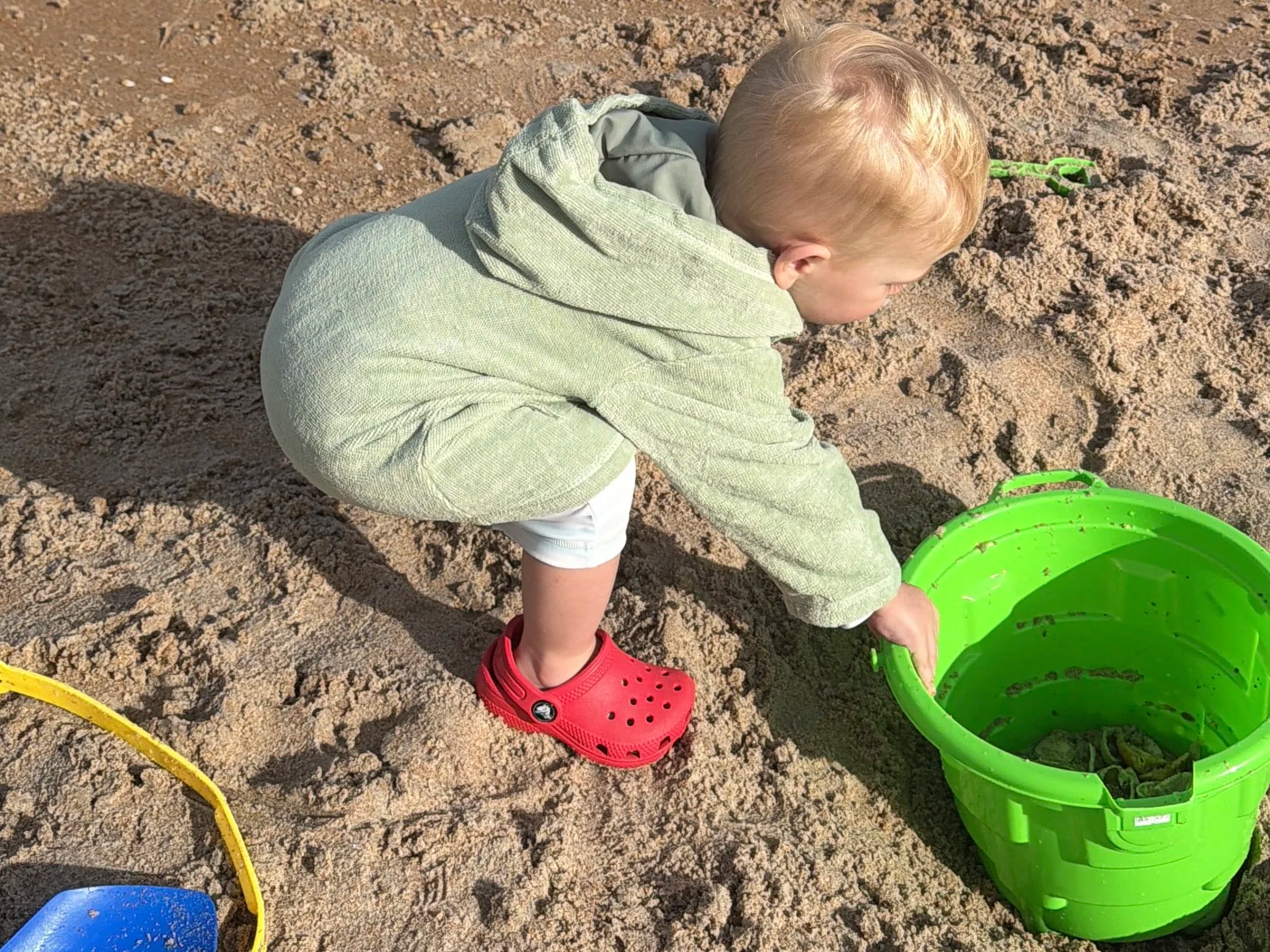 A toddler is playing on the beach. He has a green bucket inside of a hole in the sand.