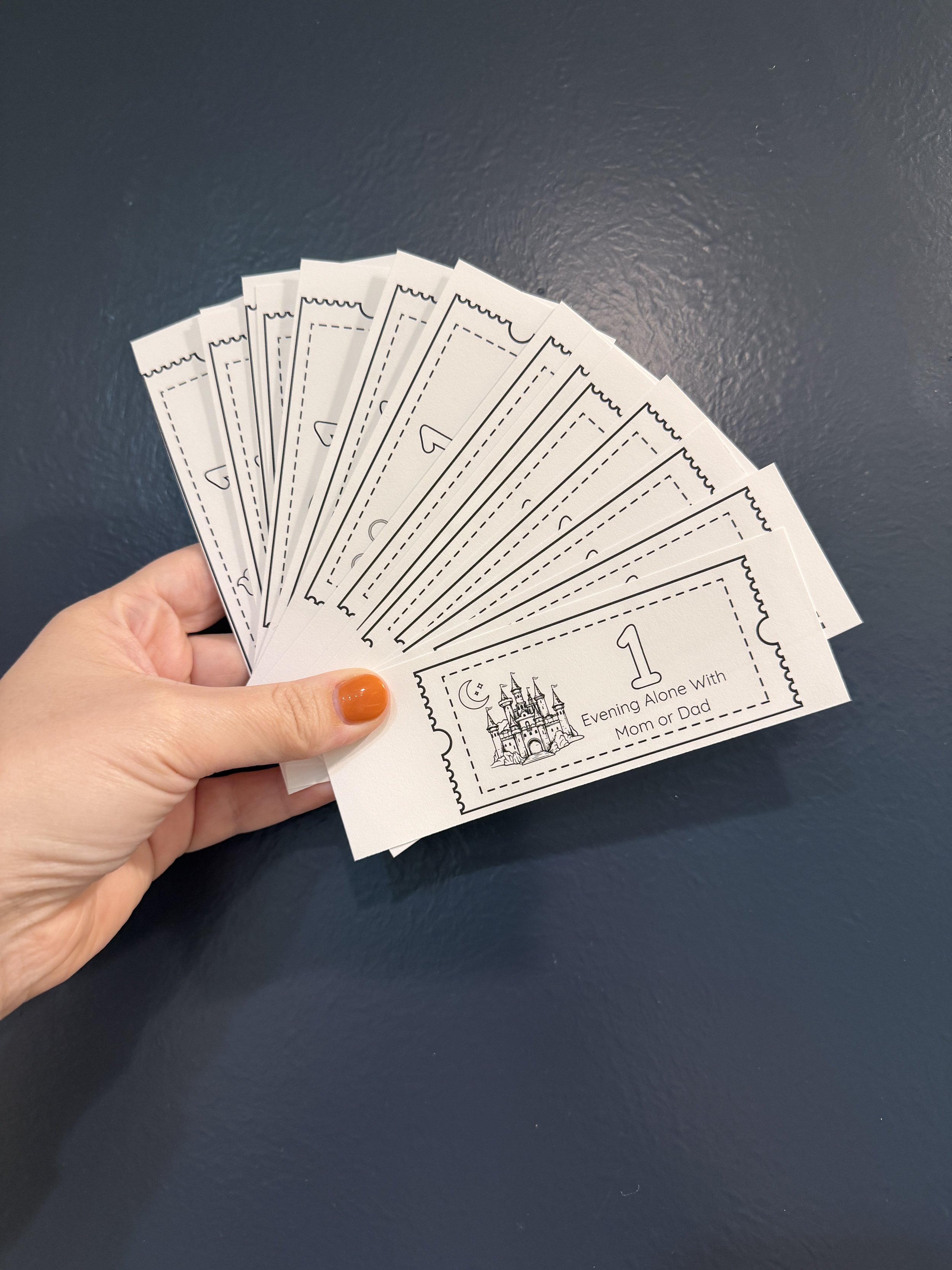 A hand with orange-painted nails holding a set of paper tickets with designs and text, including a castle and the phrase "Evening Alone With Mom or Dad."