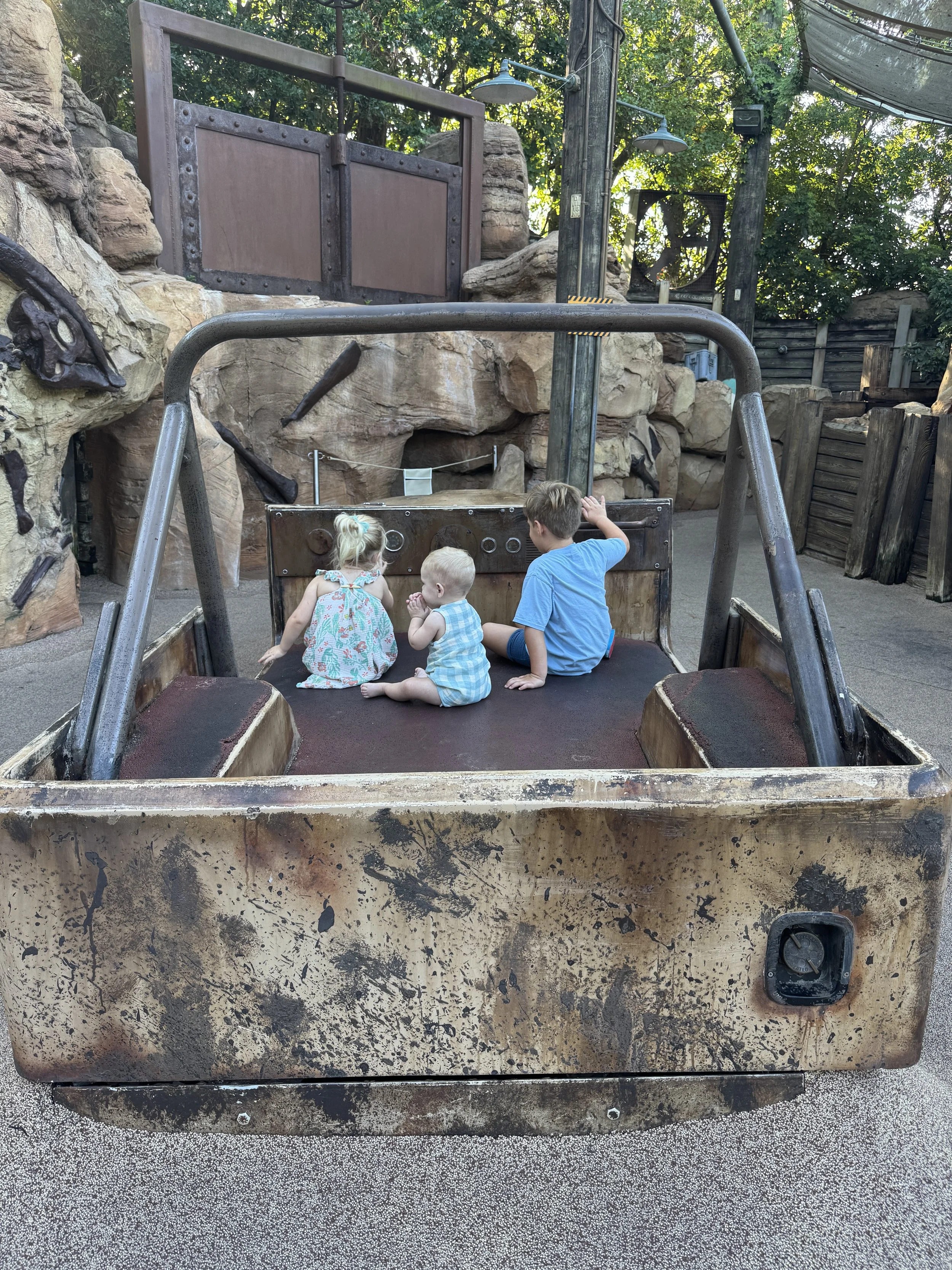 Three children sitting inside a worn, vintage-style vehicle or cart at an outdoor playground with rock formations, trees, and wooden structures in the background.