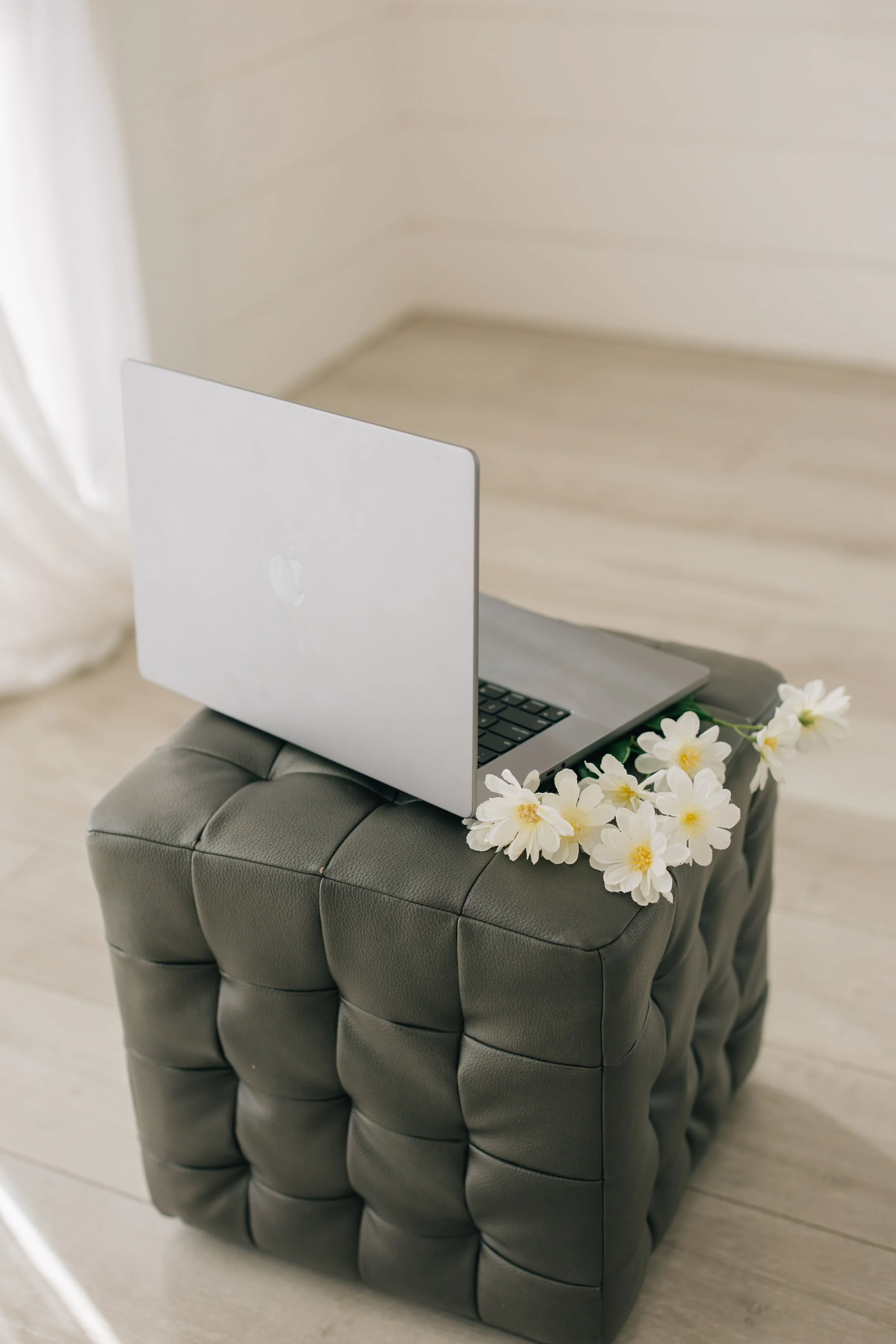 A silver MacBook laptop with a partially open lid, resting on a tufted dark gray ottoman decorated with white flowers, in a room with light-colored wooden flooring and a white wall in the background.