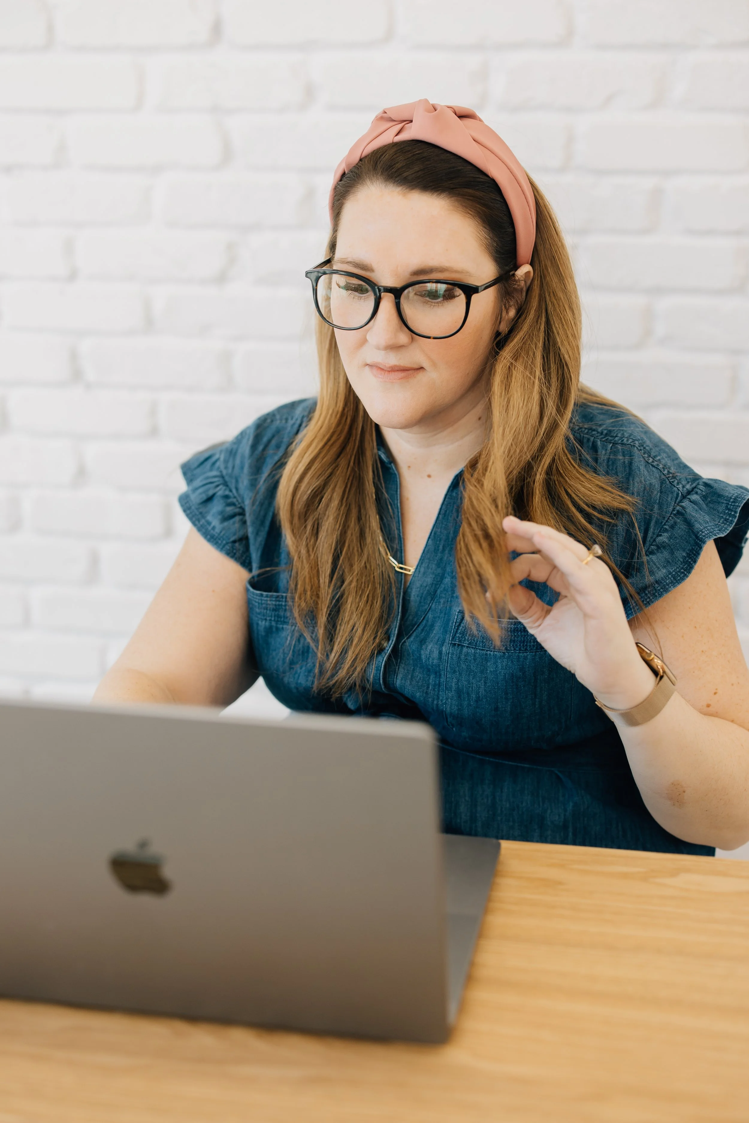 A woman with a pink headband looks down at her laptop as she touches her hair.