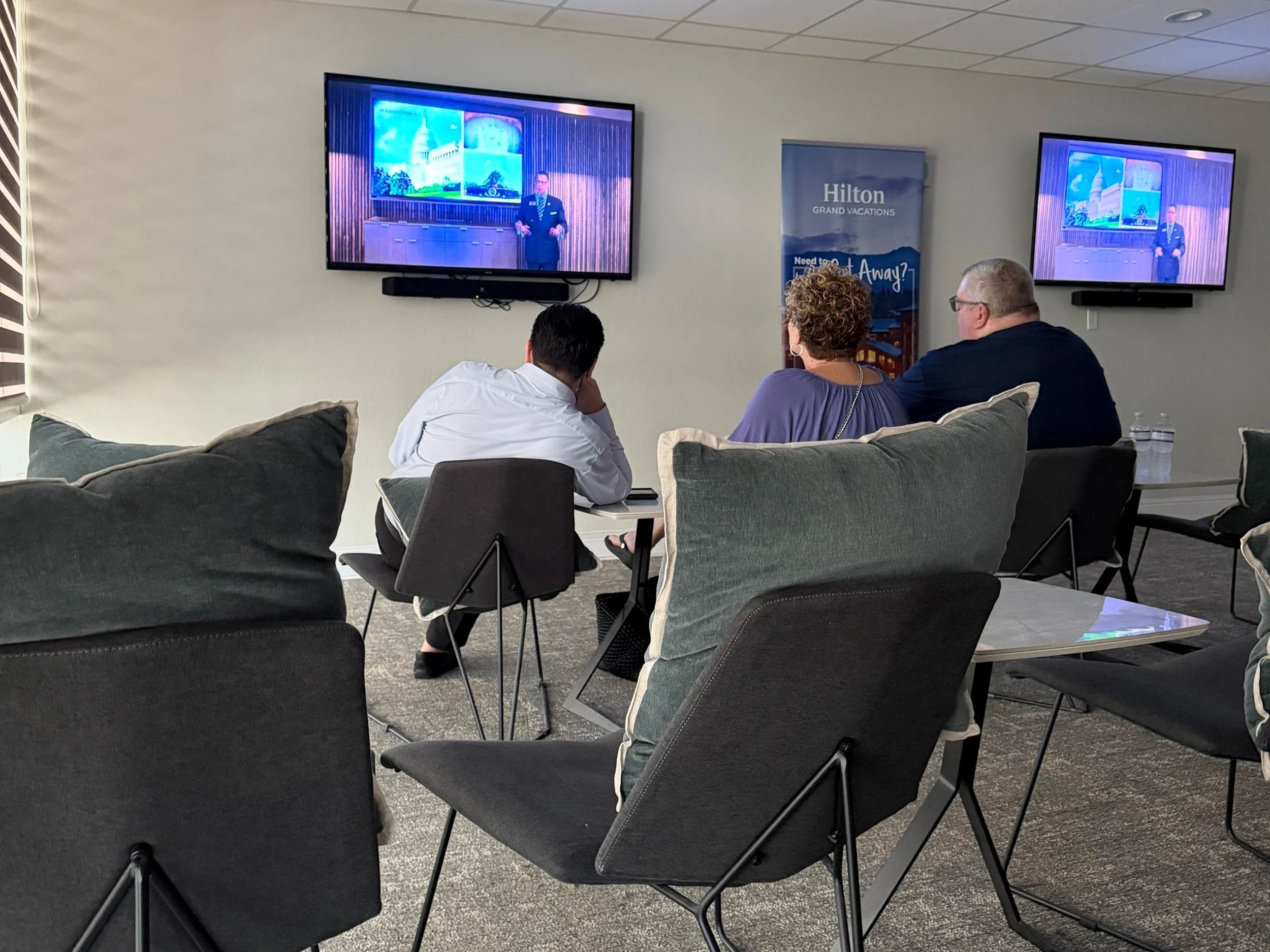 A room with grey chairs and carpet. On the wall are a few TVs showing a recording of a timeshare presentation.