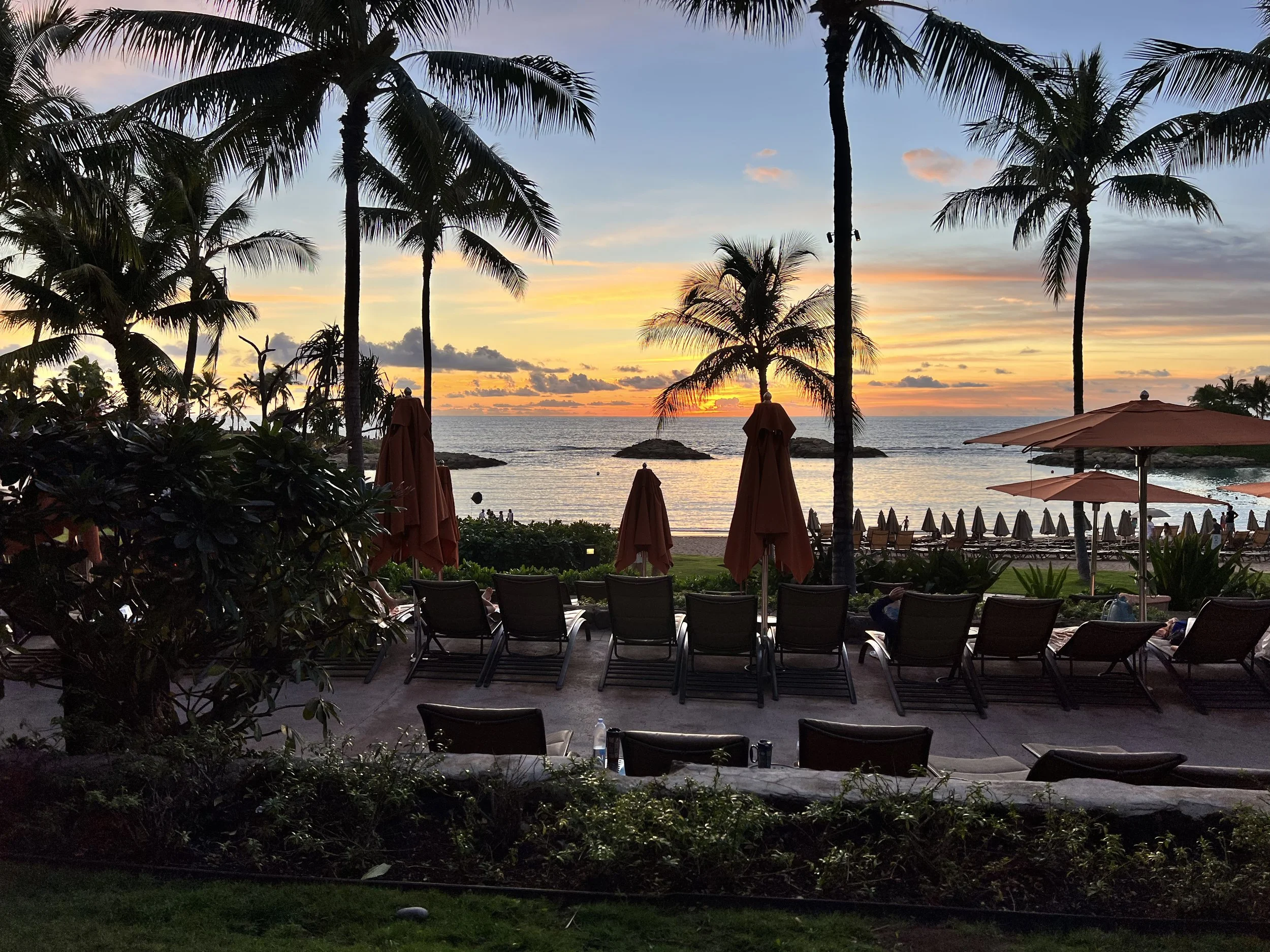 A Hawaiian beach at sunset. You can see grass, beach chairs, and the beach all in front of an orange and blue sunset sky,