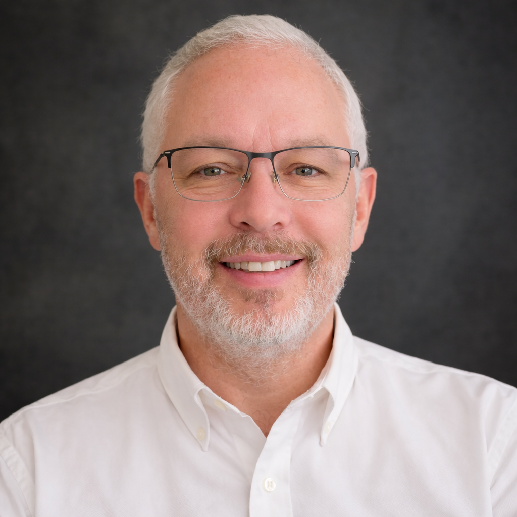 A smiling older man with glasses, a beard, and white hair, wearing a white collared shirt, against a dark background.
