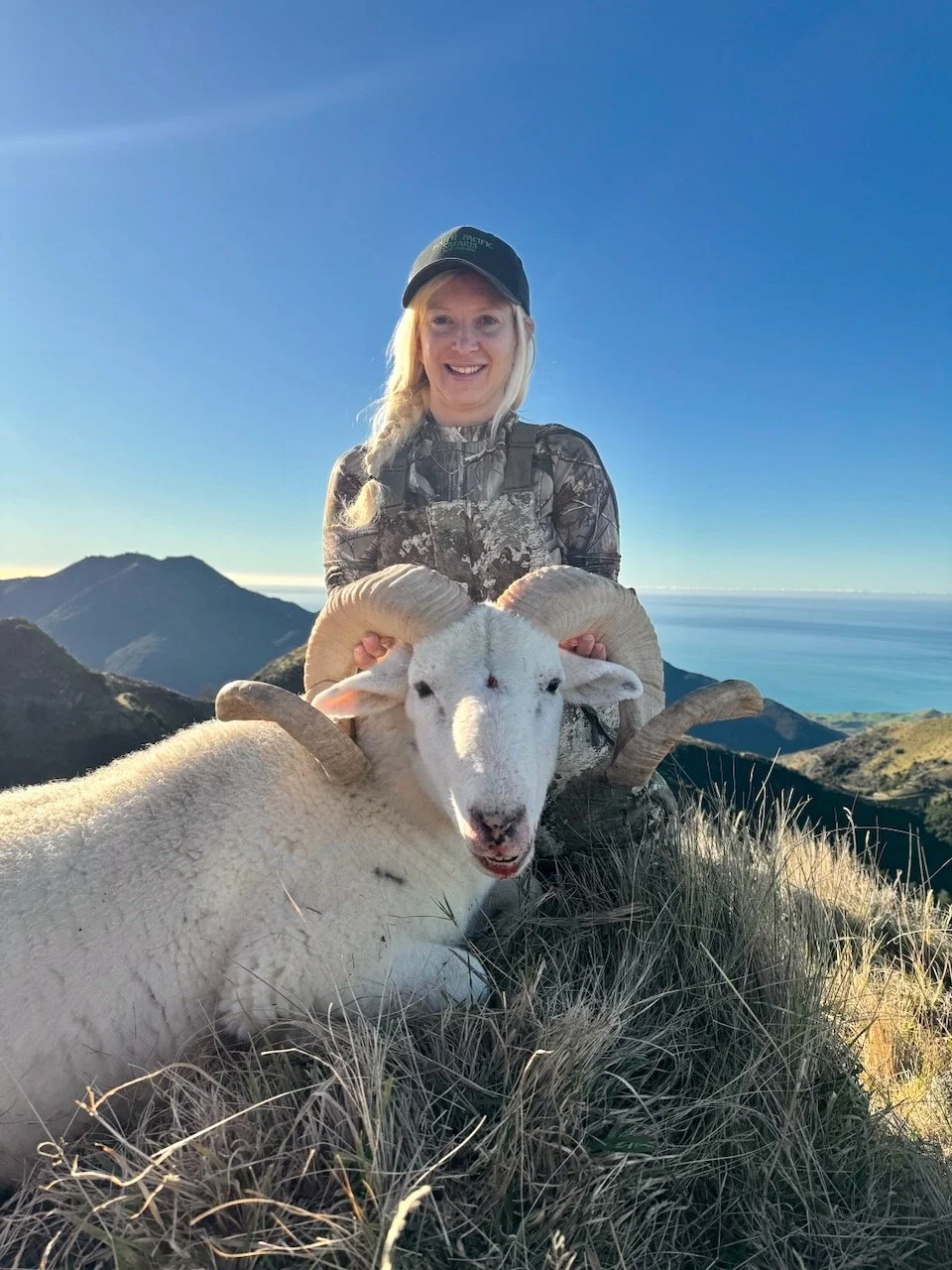A woman in outdoor camouflage clothing and a hat is smiling while holding a mountain goat with curled horns, on a mountain with a scenic background of blue sky, peaks, and ocean.