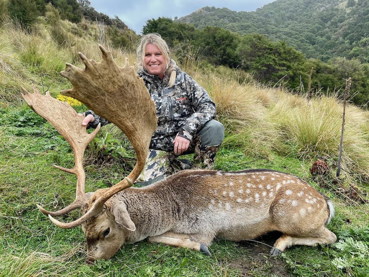A woman in camouflage clothing kneels on a grassy hillside, smiling, after hunting a large deer with impressive antlers, which is lying on the grass beside her with its head resting on the ground.