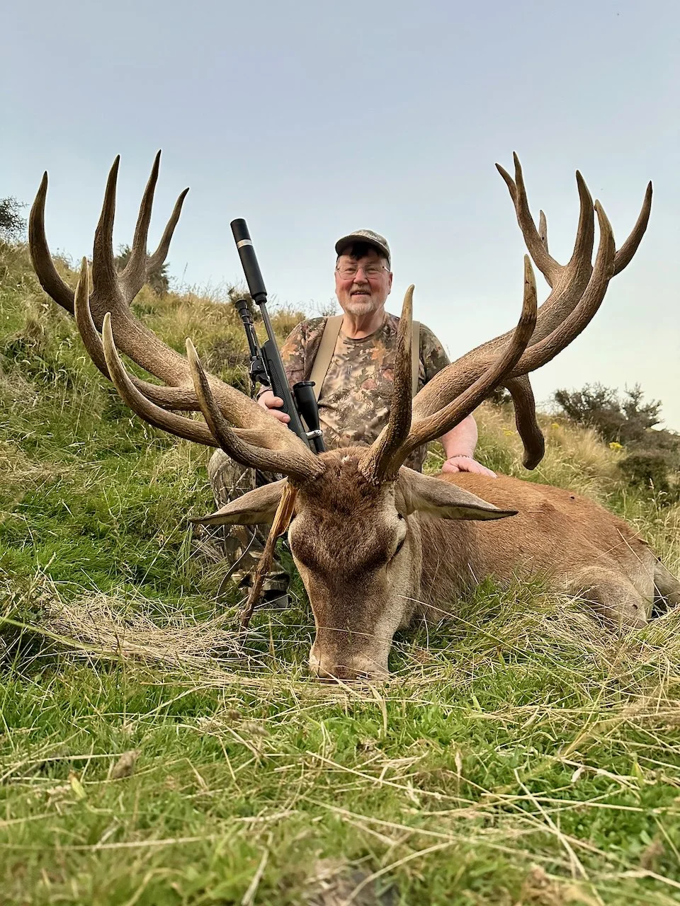 A man in camouflage clothing sitting behind a large dead deer with big antlers, holding a rifle, on a grassy hillside.