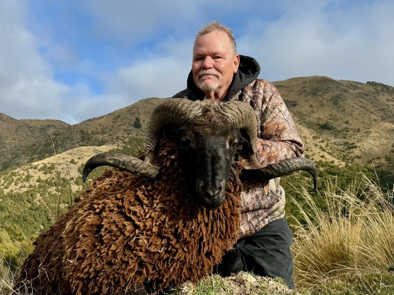 A man in camouflage clothing kneels behind a large ram with curved horns, outdoors in a mountainous area with grass and bushes, under a partly cloudy sky.