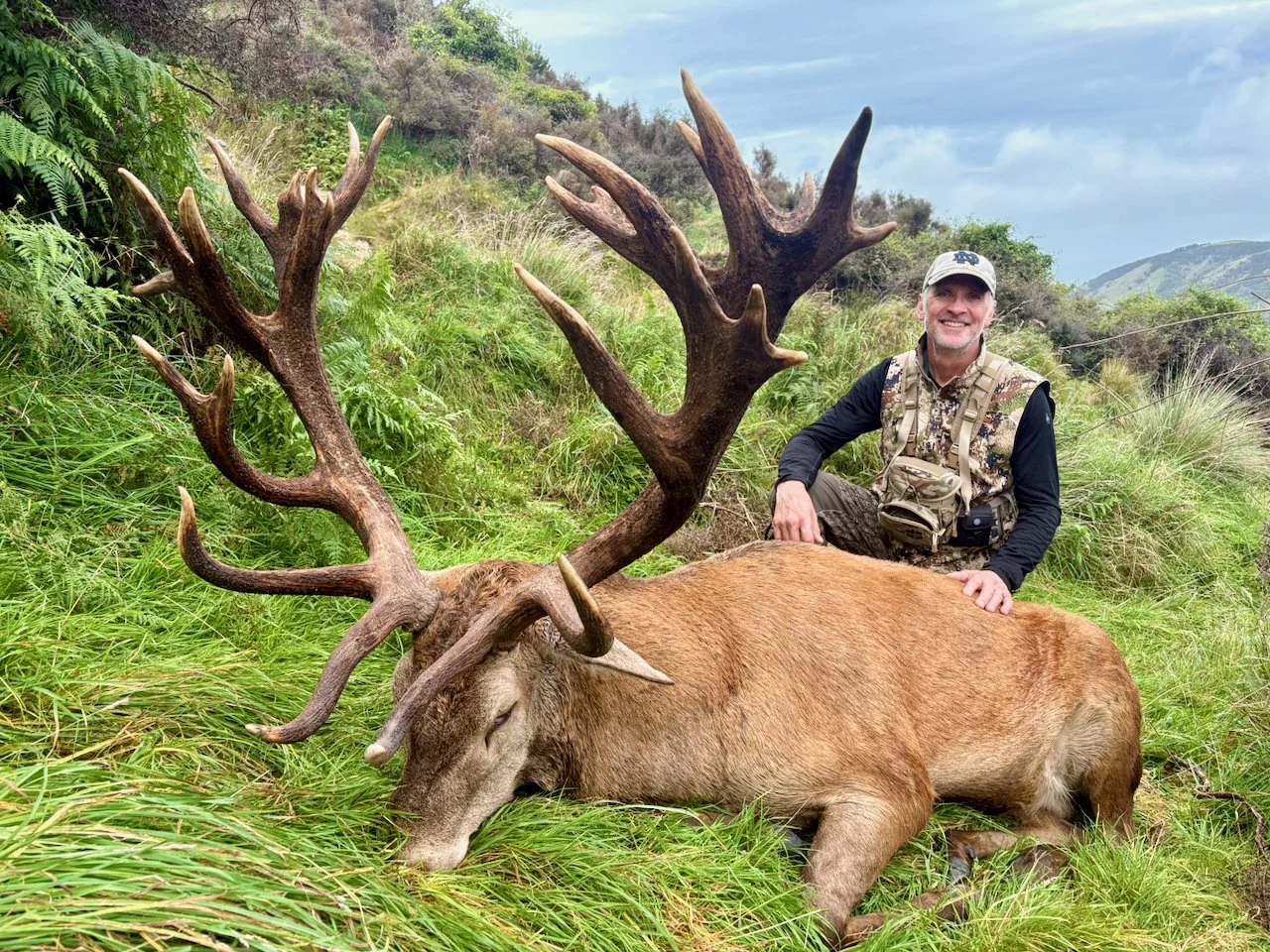 A man in camouflage clothing and a cap kneels next to a large dead elk with impressive antlers, lying on grass in a hilly, green landscape with cloudy skies.