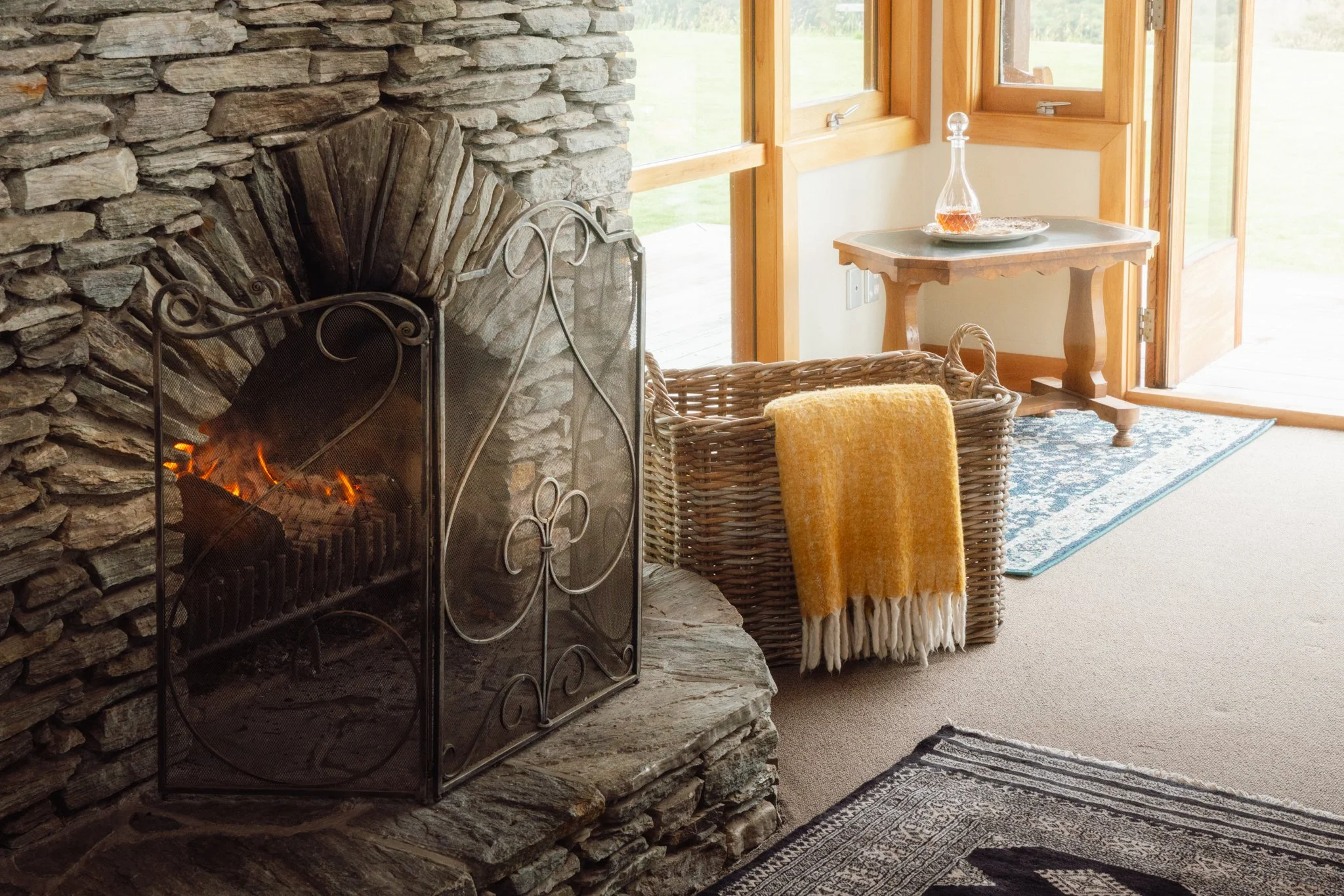 A cozy living room corner with a stone fireplace, a wicker chair with a yellow blanket, a small wooden table with a glass decanter, and a patterned rug. Large windows reveal a green outdoor landscape.
