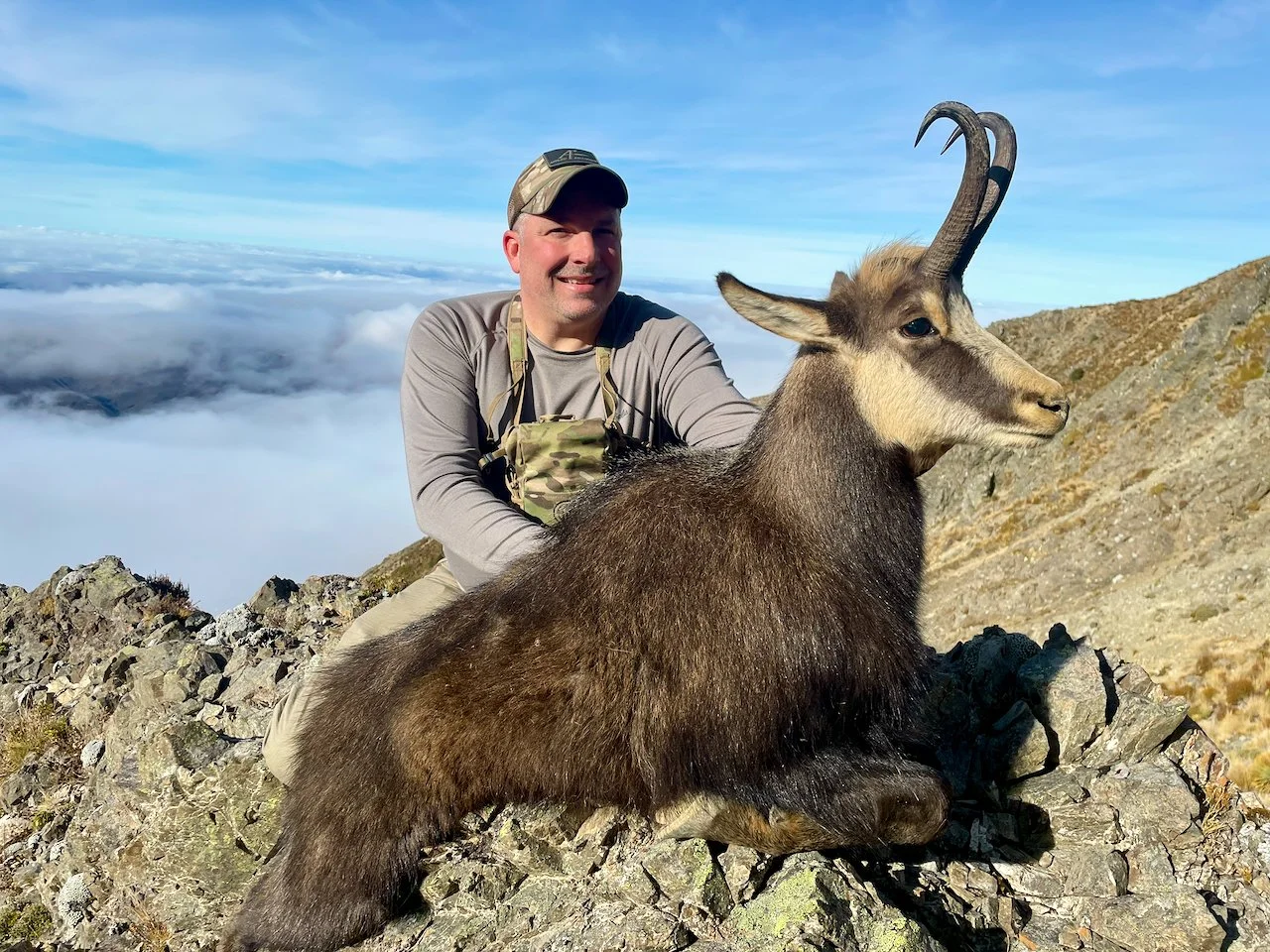 A man in outdoor gear sitting on a mountain, holding a mountain goat with curved horns, with clouds and mountain peaks in the background.