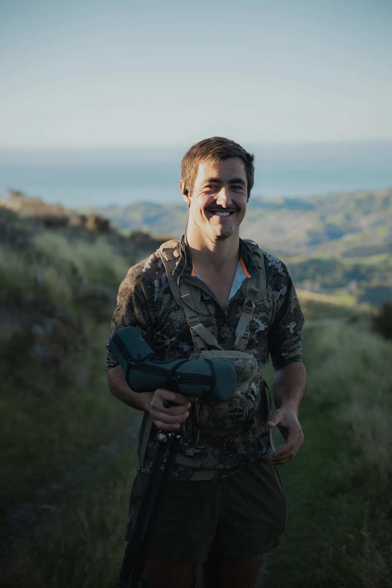 A man in outdoor camouflage clothing smiling while standing on a grassy trail with rolling hills and the ocean in the background.