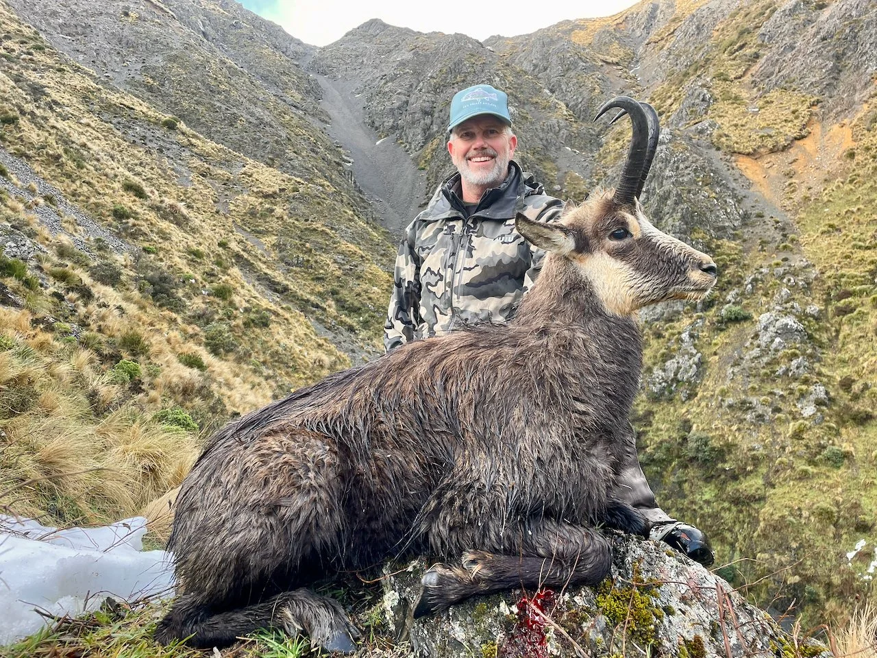 A smiling man in camouflage jacket and blue cap posing with a harvested mountain goat in a rugged, grassy, mountainous landscape.