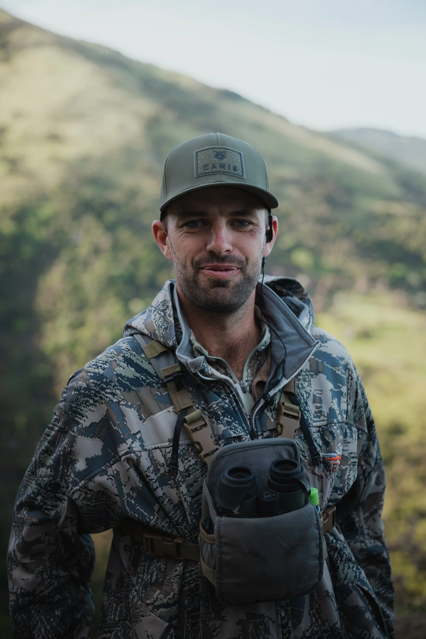 A man wearing a gray cap with and a camouflage jacket, fishing in a natural outdoor setting with mountains and greenery in the background.