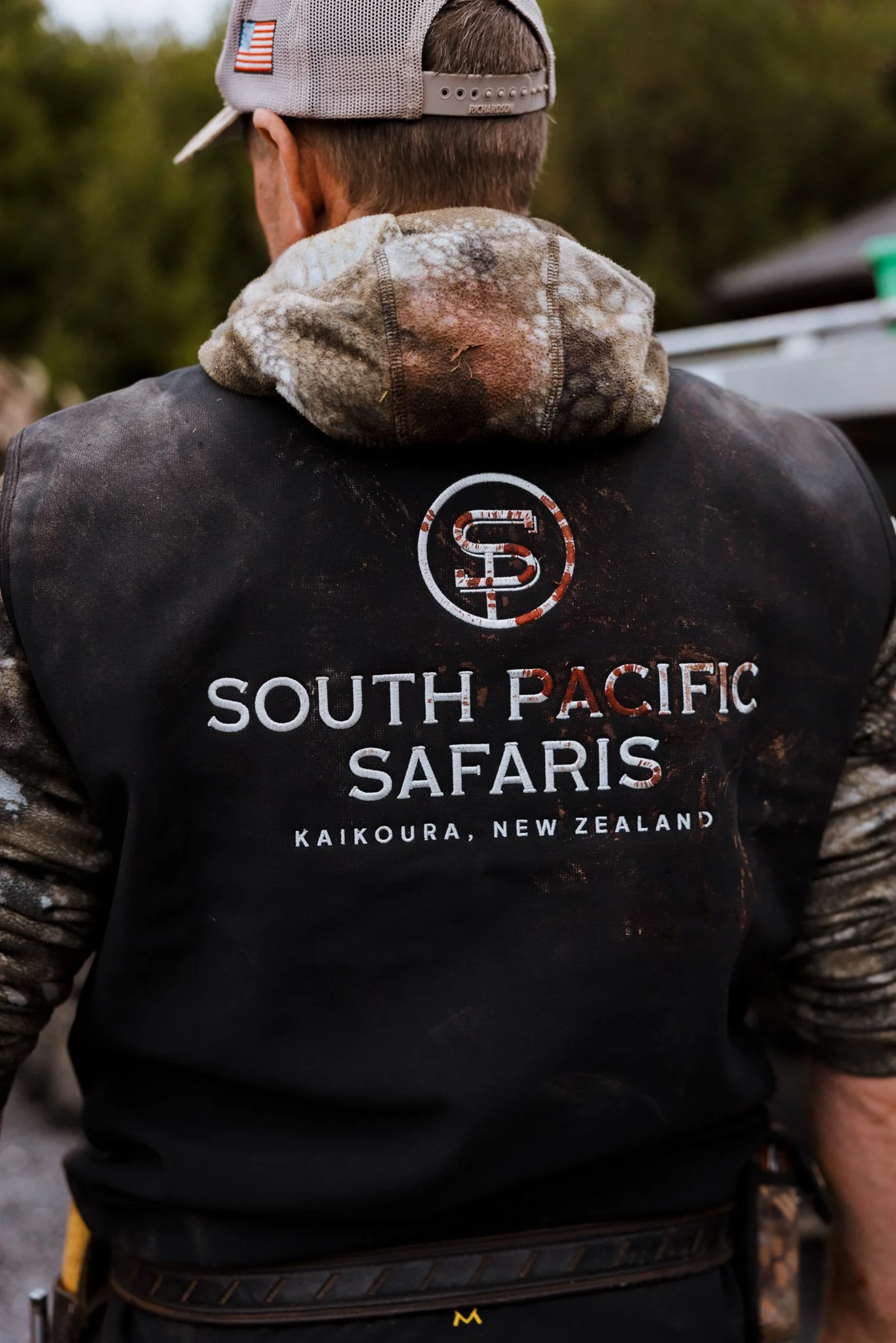 Back of a person wearing a baseball cap and a black South Pacific Safaris jacket, with a camouflage hoodie, standing outdoors.
