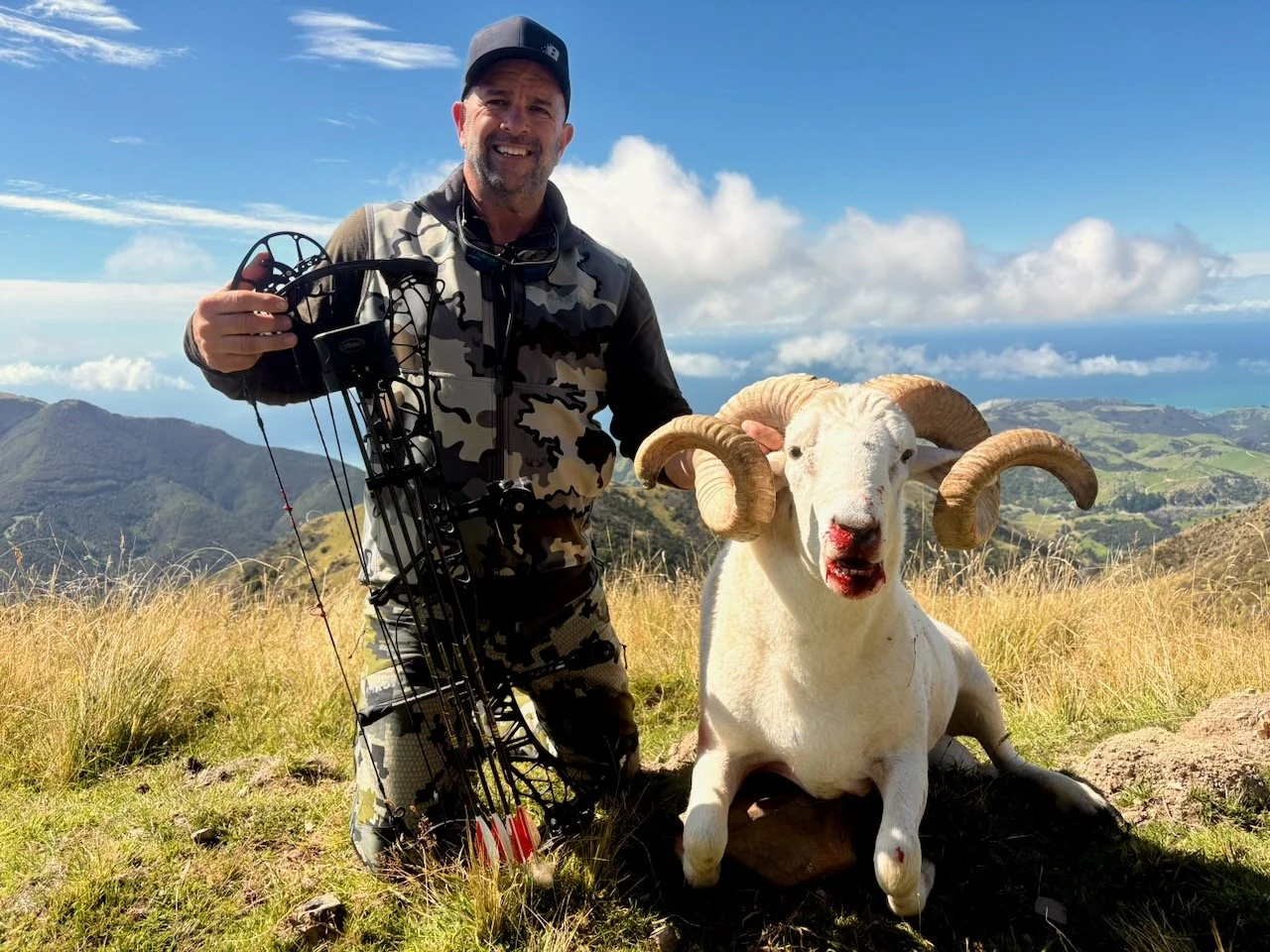 A man smiling and holding a bow stands next to a white mountain goat with large curved horns, on a grassy hillside with mountains and the ocean in the background under a partly cloudy sky.