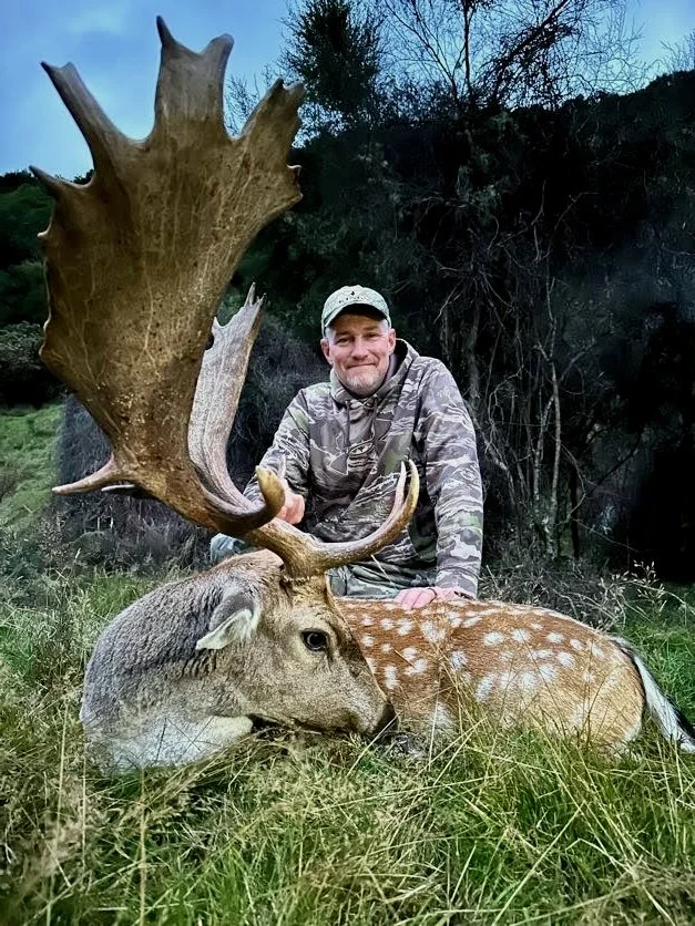 A man in camouflage clothing and a cap kneeling behind a large fallen deer with impressive antlers, in a grassy outdoor setting with trees and a clear sky in the background.