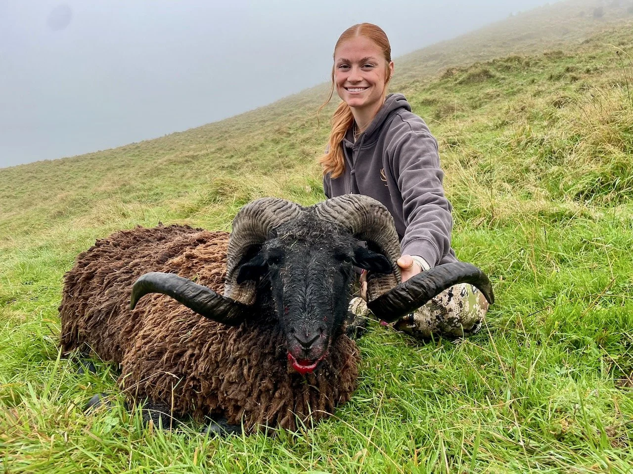 A smiling woman in a gray hoodie kneels next to a large ram with curled horns on a grassy hillside.
