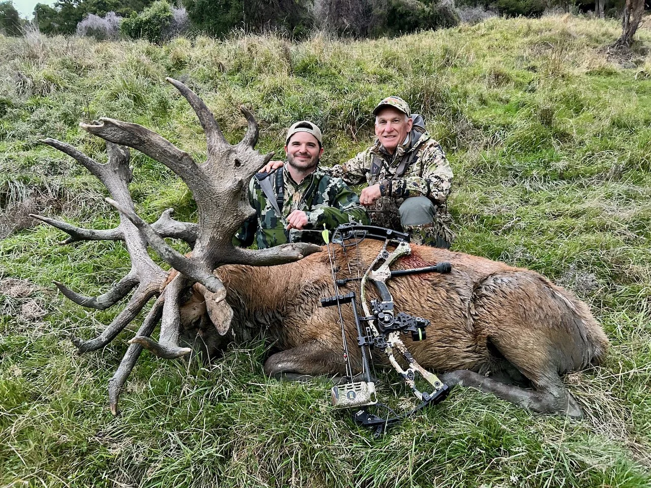 Two hunters posing with a large, freshly killed elk with impressive antlers, lying on its side in a grassy field, with a bow and arrows resting on the elk's body.
