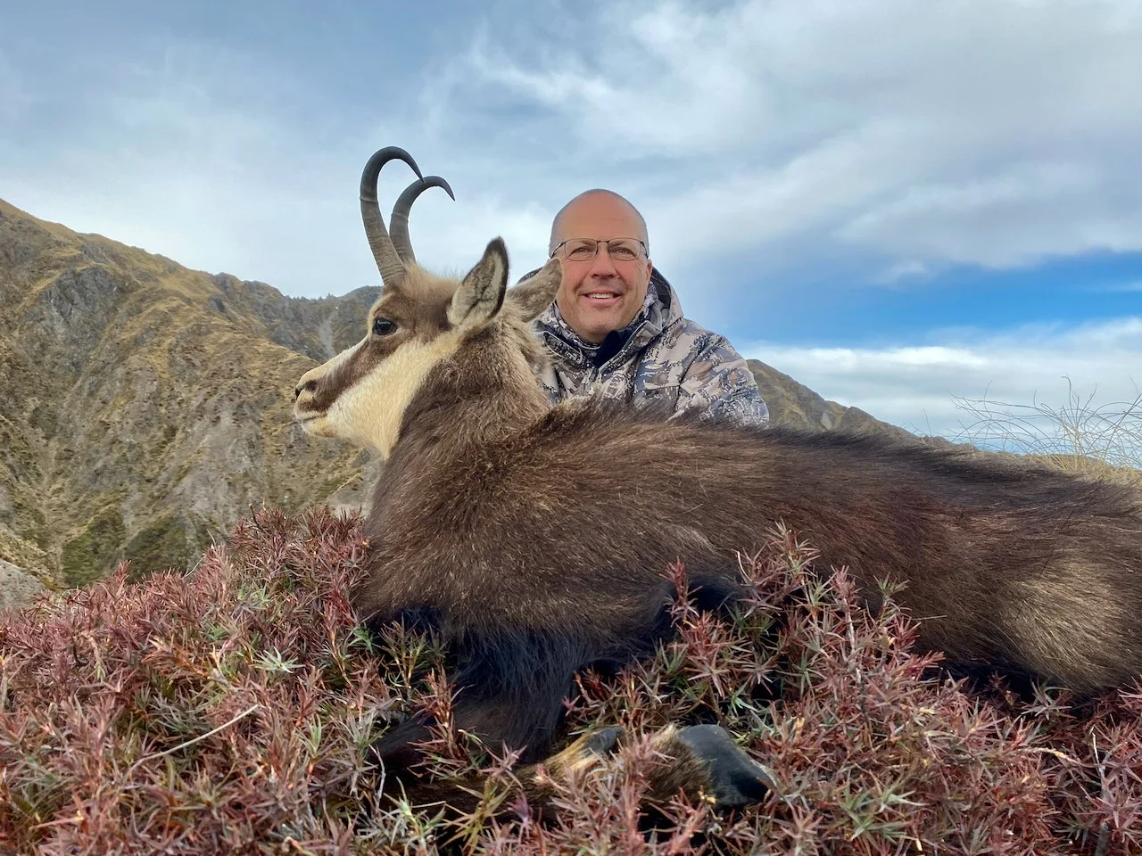 A man wearing glasses and camouflage jacket smiling while sitting behind a mountain goat with curved horns in a mountainous landscape with cloudy sky.
