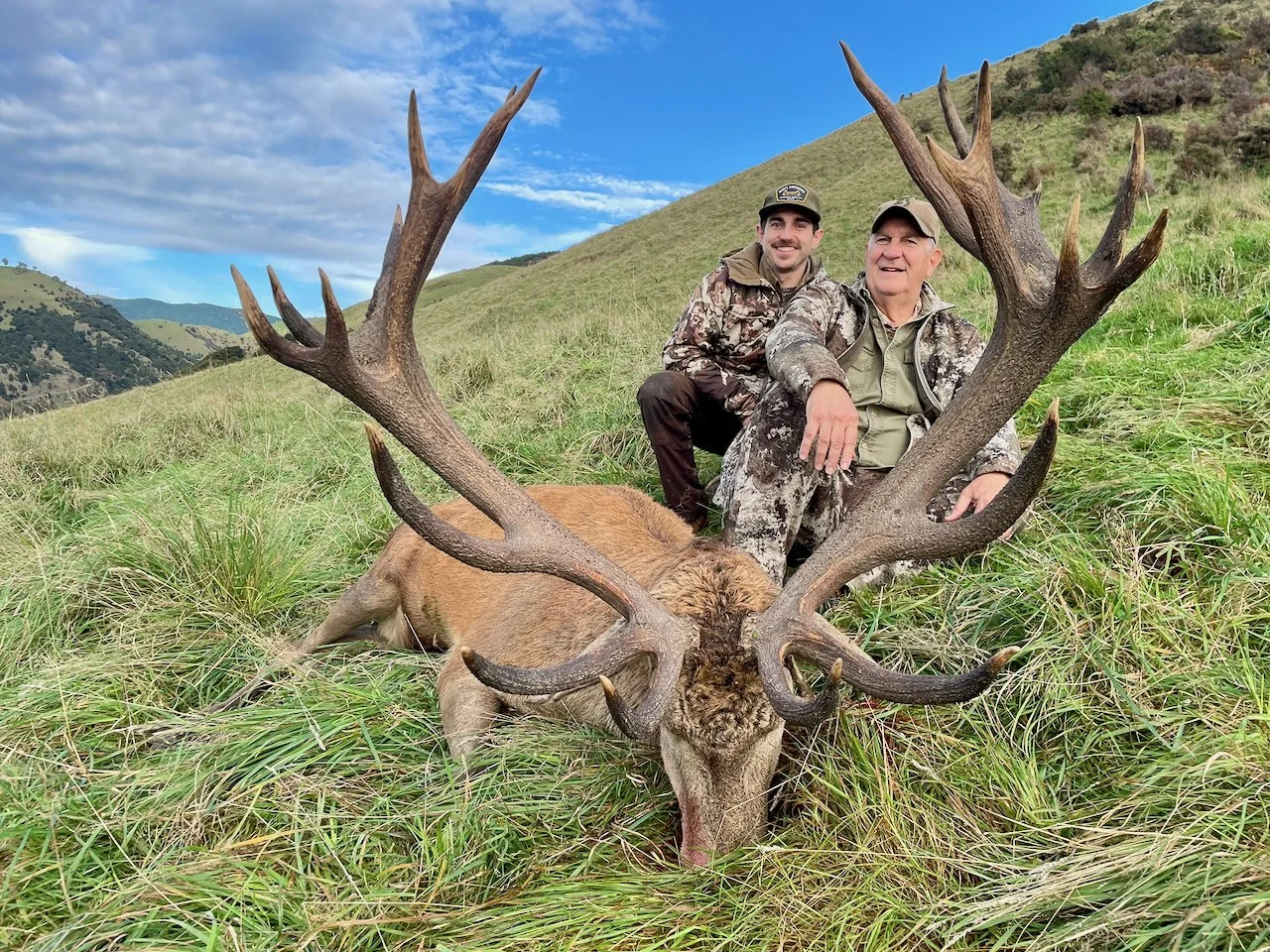 Two hunters posing with a large deer with antlers in a grassy field on a hillside, mountains and partly cloudy sky in the background.