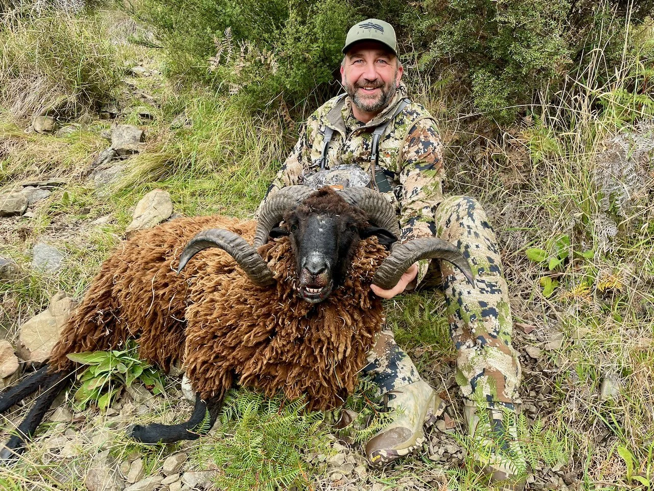 A man in camouflage outdoor clothing and a cap smiling while holding a large, dark-furred sheep with twisted horns in a natural, grassy, rocky area.