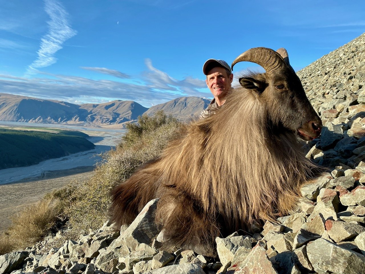 A man wearing a baseball cap sitting next to a large wild goat with thick fur and large curved horns on a rocky hillside, with mountains and a valley in the background under a blue sky with clouds.
