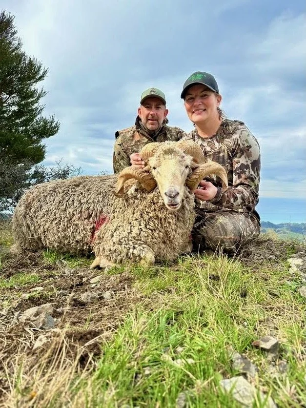 A man and woman in camouflage clothing kneel on the ground behind a large sheep with impressive horns, which they appear to have hunted, as indicated by the blood on its side. They are outdoors, with a cloudy sky and some trees in the background.